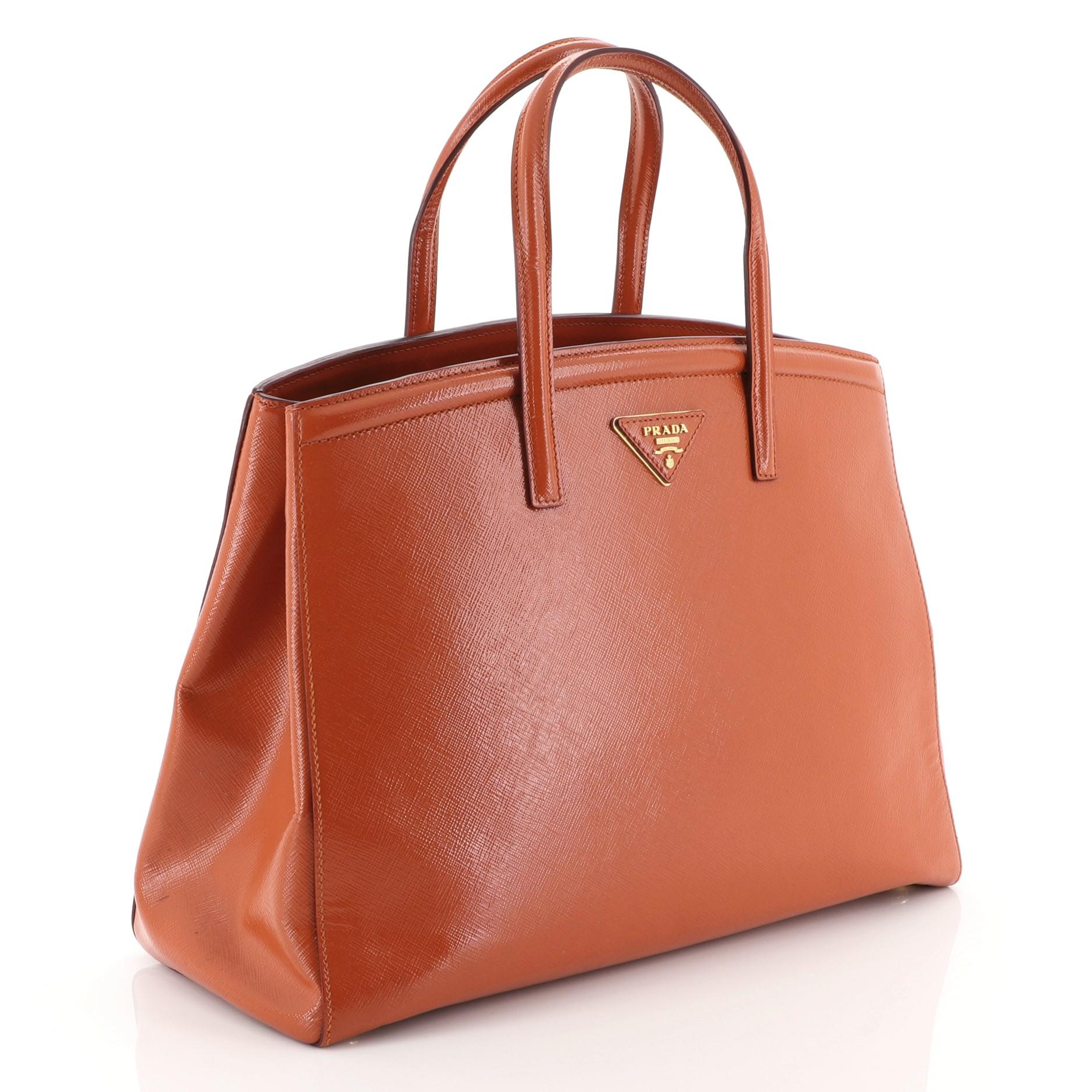 This Prada Slim Convertible Tote Vernice Saffiano Leather East West, crafted in orange vernice saffiano leather, features dual top handles and gold-tone hardware. It opens to an orange fabric interior. 

Condition: Great. Creasing on sides, minor
