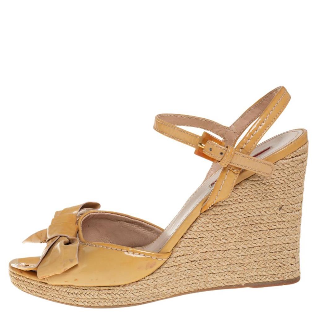 These laid-back sandals by Prada are made from beige patent leather and raffia. They are accented with bow detailing with the brand name and ankle straps with buckle fastenings. They are equipped with 11 cm wedge heels and leather-lined
