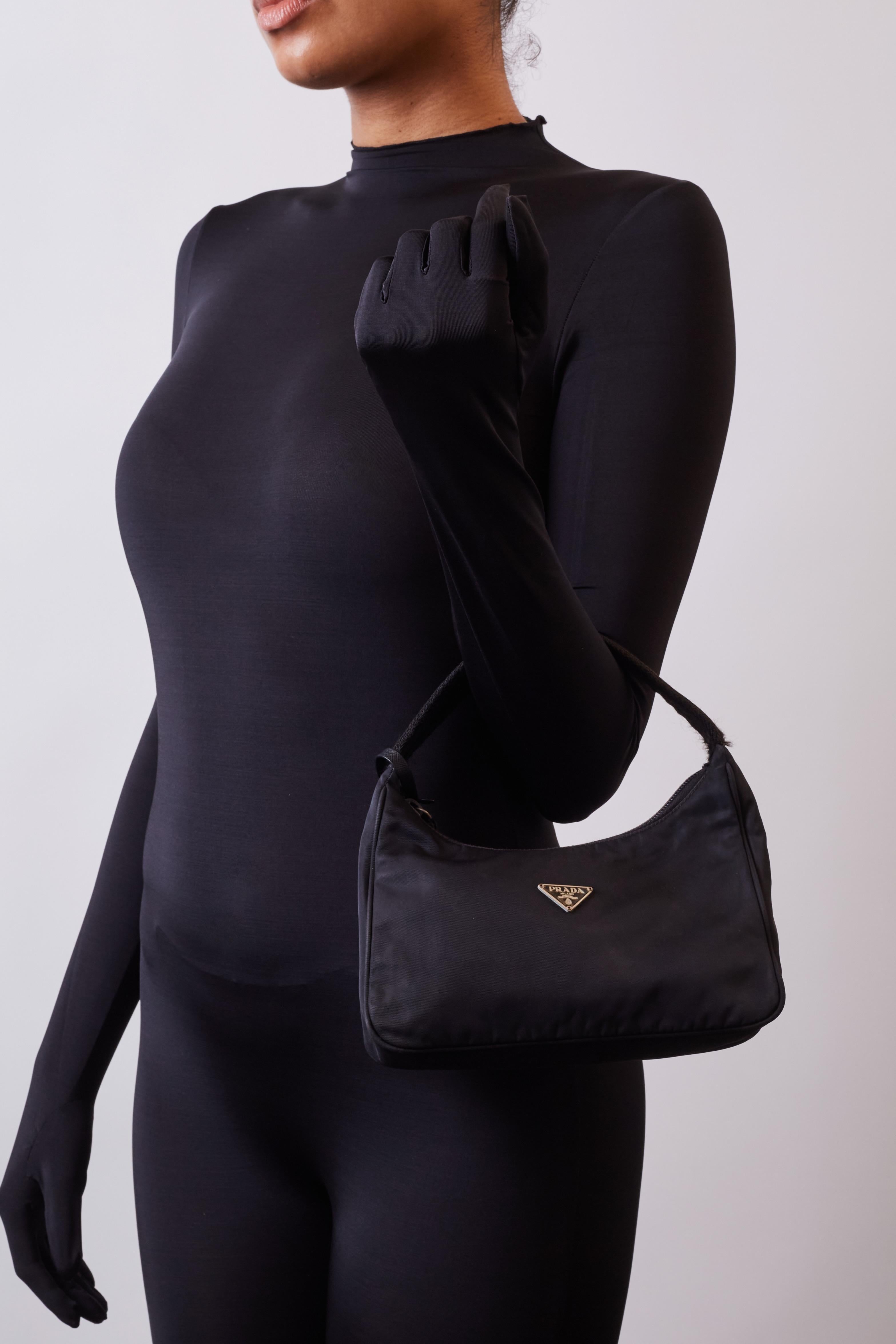 This bag tote is made of black nylon fabric. The shoulder bag features a polished silver tone enameled black Prada logo on the front, a tall black durable nylon shoulder strap, and a broad zipper top opening. The top zipper opens to a matching nylon