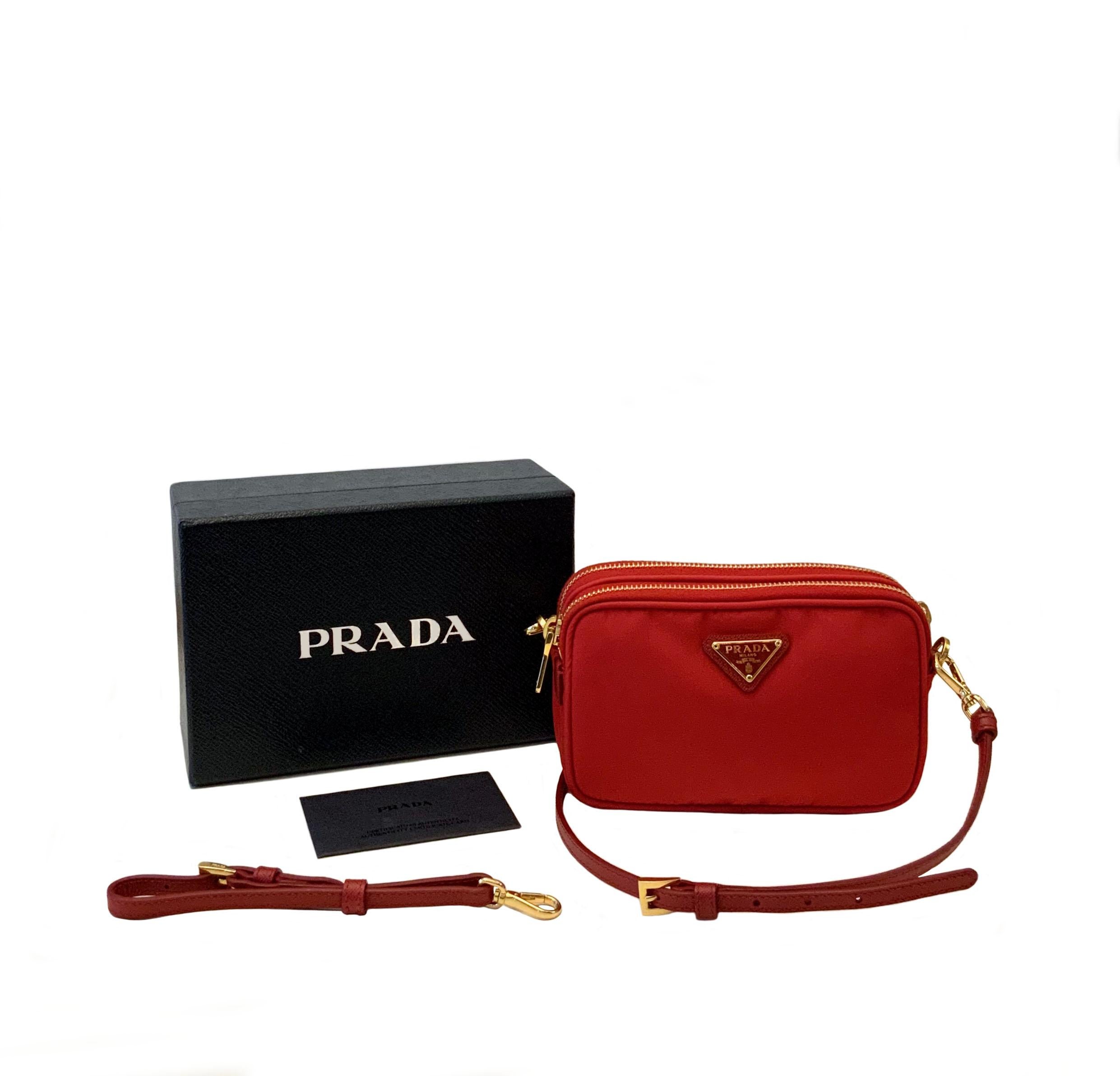 Este bolso bandolera de segunda mano pero nuevo está fabricado en nailon Prada de color rojo. 
El bolso tiene una larga correa cruzada de piel y un asa sencilla de piel adicional y extraíble.
Dos cremalleras superiores se abren a un interior con el