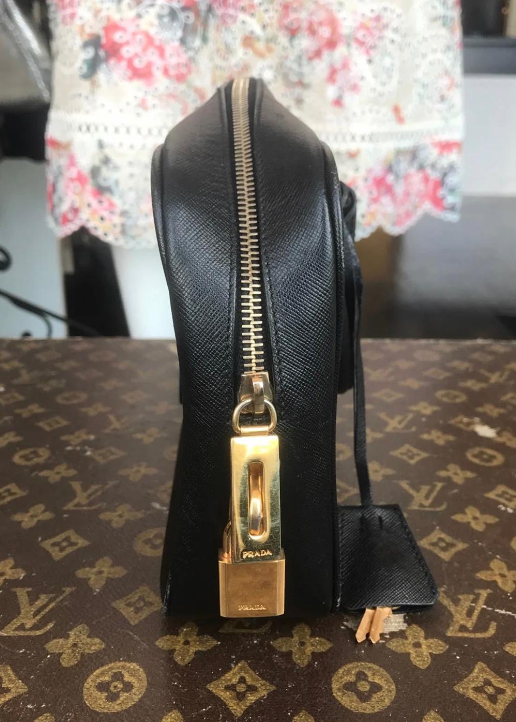 Black grained leather. Gold-tone hardware. Dual top handles. Prada embossed logo on front. Zippered closure at top. Protective feet at bottom. Lock and two keys. One interior zippered pocket.