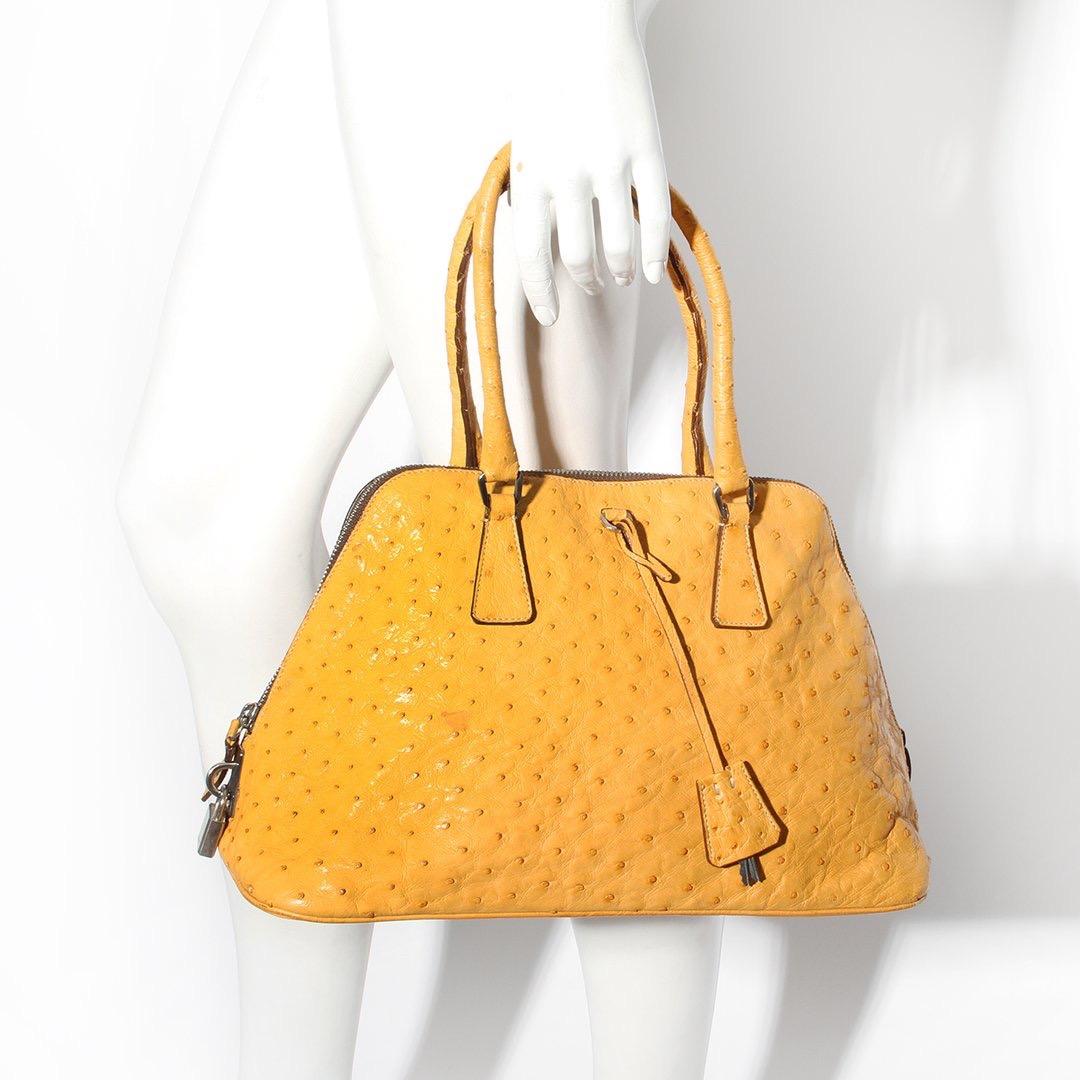 Prada Leather Trapazoid Handbag 
Made in Italy 
Mustard yellow color
Ostrich leather 
Small double handles at top of tote 
Flat bottom 
Silver tone lock hardaware
Lock is stamped Prada 
Attached silver tone keys for lock 
Lined with green Prada logo
