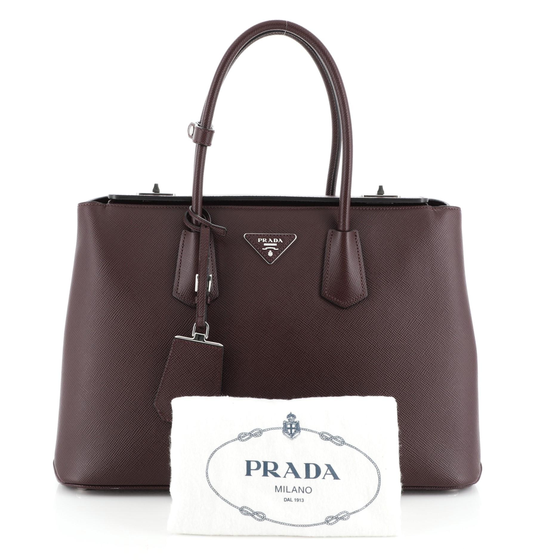 This Prada Turnlock Cuir Twin Tote Saffiano Leather Medium, crafted in red saffiano leather, features dual rolled leather handles, protective base studs, and silver-tone hardware. Its dual turn-lock closure opens to a black and red leather interior