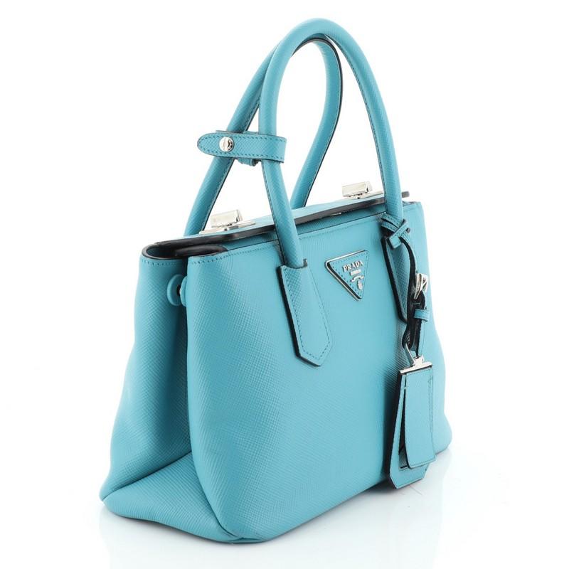 This Prada Turnlock Cuir Twin Tote Saffiano Leather Mini, crafted from blue saffiano leather, features dual-rolled leather handles, angular silhouette, inverted triangle Prada logo, side snap buttons, and silver-tone hardware. Its fold-over top with