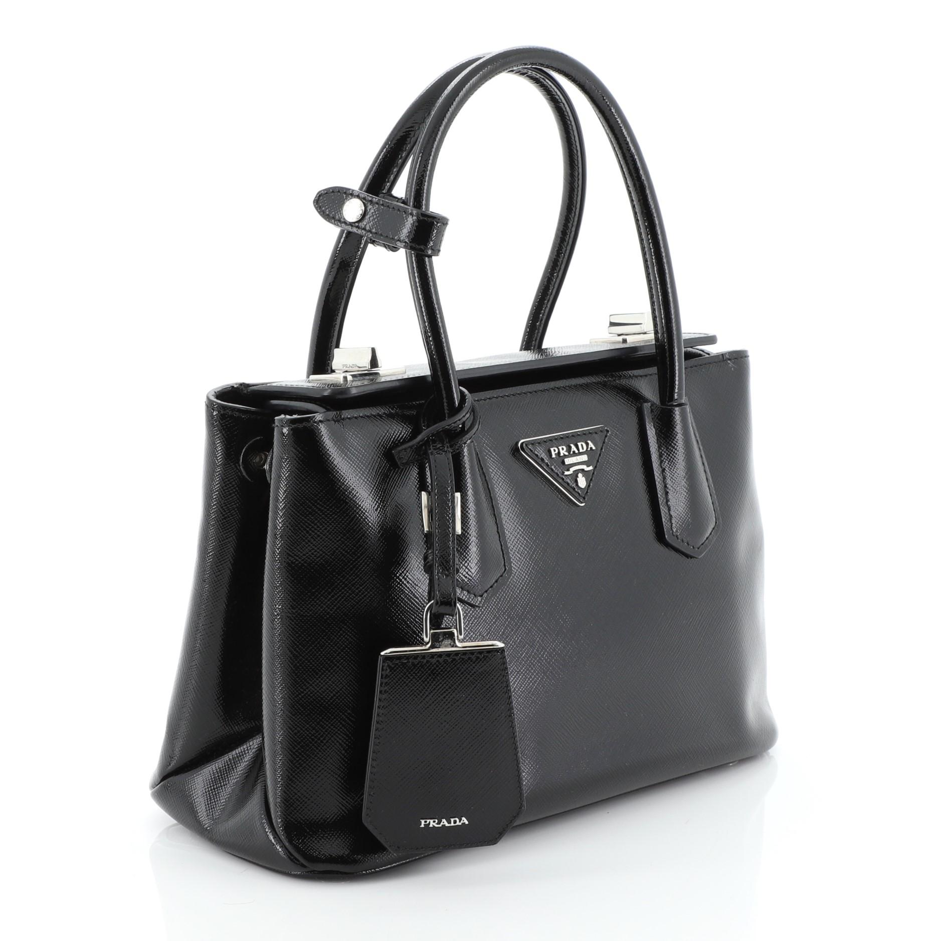 This Prada Turnlock Cuir Twin Tote Vernice Saffiano Leather Mini, crafted in black vernice saffiano leather, features dual rolled leather handles, protective base studs, and silver-tone hardware. Its dual turn-lock closure opens to a red leather