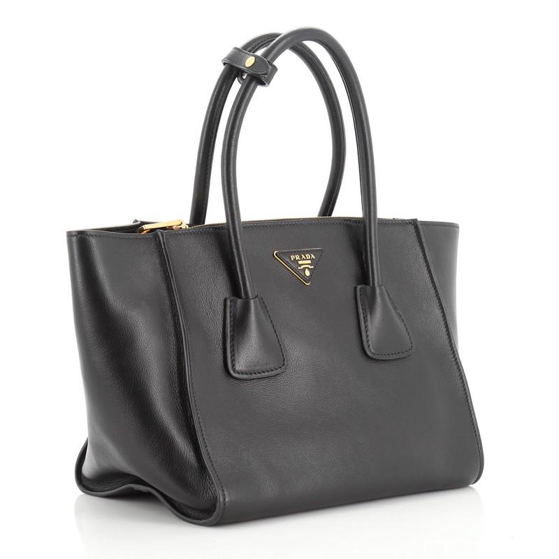 This Prada Twin Pocket Tote City Calfskin Small, crafted from black calfskin leather, features dual rolled top handles, Prada triangle logo, and gold-tone hardware. It opens to a black fabric interior with zip and slip pockets. 

Estimated Retail