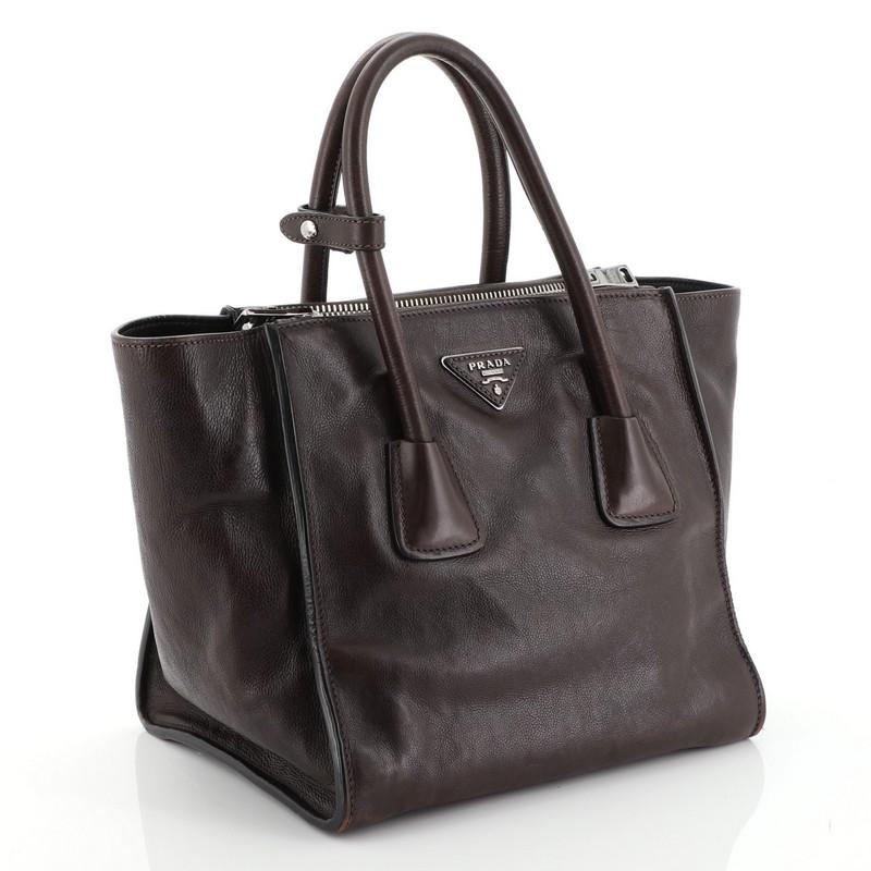 This Prada Twin Pocket Tote Glace Calf Small, crafted from brown glace calf leather, features tall dual-rolled top handles, signature center Prada triangle logo, protective base studs and silver-tone hardware. Its snap closure opens to a black