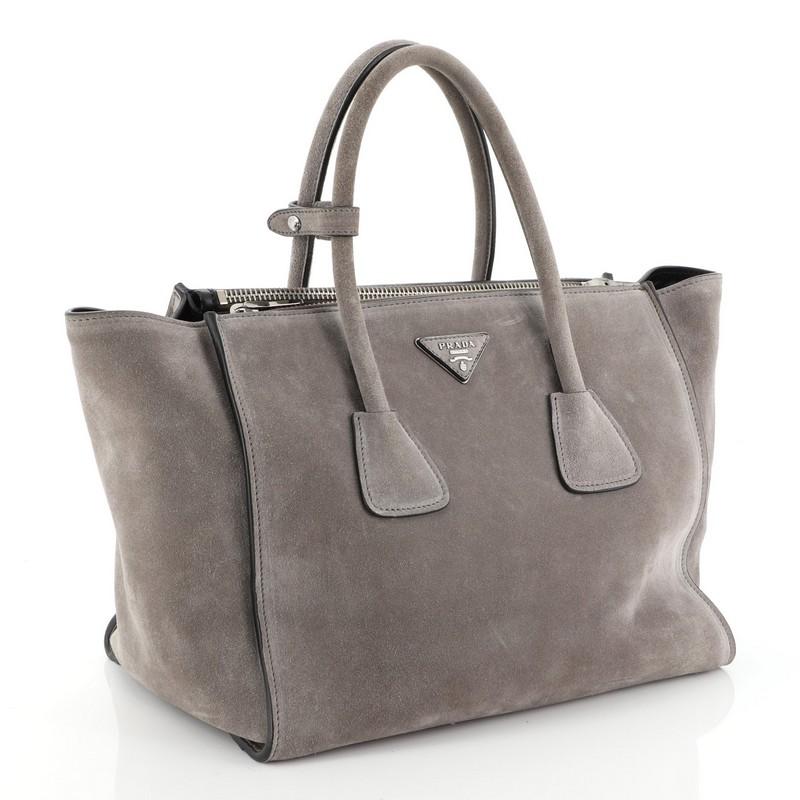 This Prada Twin Pocket Tote Suede Medium, crafted from gray suede, features tall dual-rolled handles, expanded winged sides, raised Prada logo at the center, protective base studs and silver-tone hardware accents. Its wide open top and snap closure