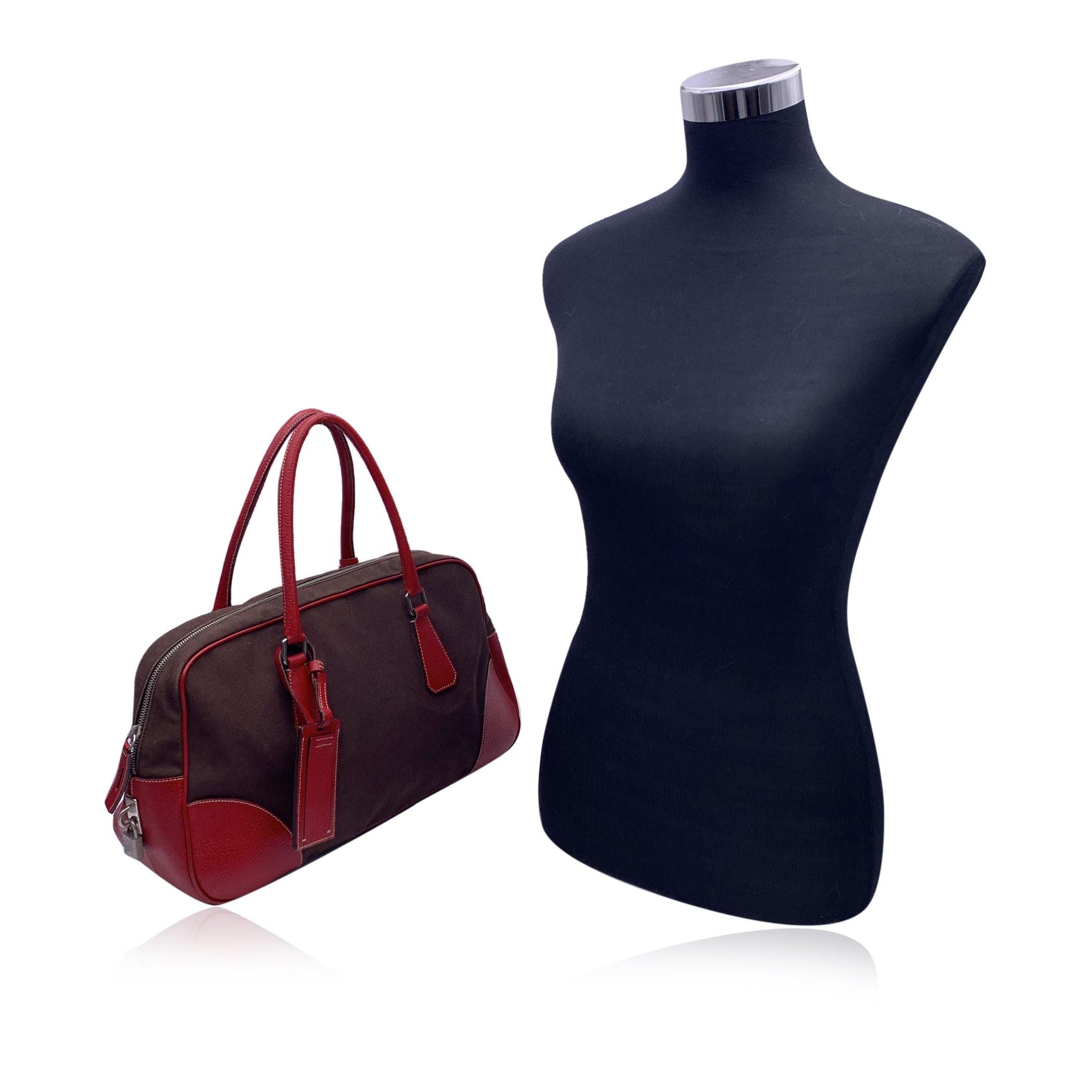 Step into timeless elegance with this classic PRADA Bowling Bag, a perfect blend of luxury and functionality. Crafted from rich brown Canapa canvas, this bag is beautifully complemented by bold red leather trim and rolled top handles, offering a pop