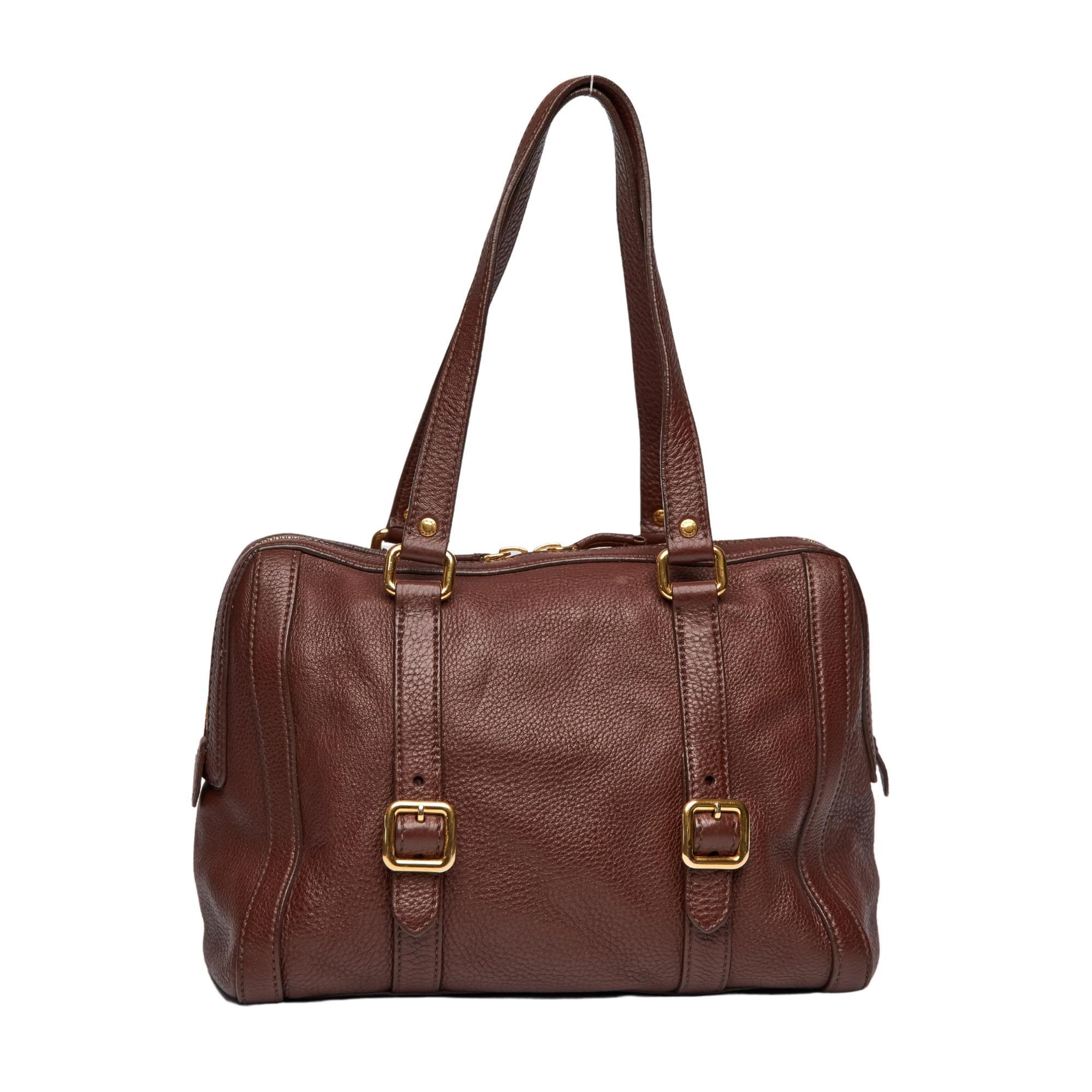 This shoulder bag is created out of fine daino deerskin leather in bruciato. The bag features long dual flat leather shoulder straps with side buckles in gold hardware. The top zips open to a brown Prada logo jacquard weave fabric with a zippered