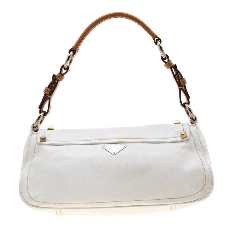 Adorned in an understated white hue, this Prada shoulder bag is beautiful. It is designed with perceptible stitching and a front flap which comes decorated with a silver-tone D-ring and bold gold and silver studs. A tan leather shoulder strap and a