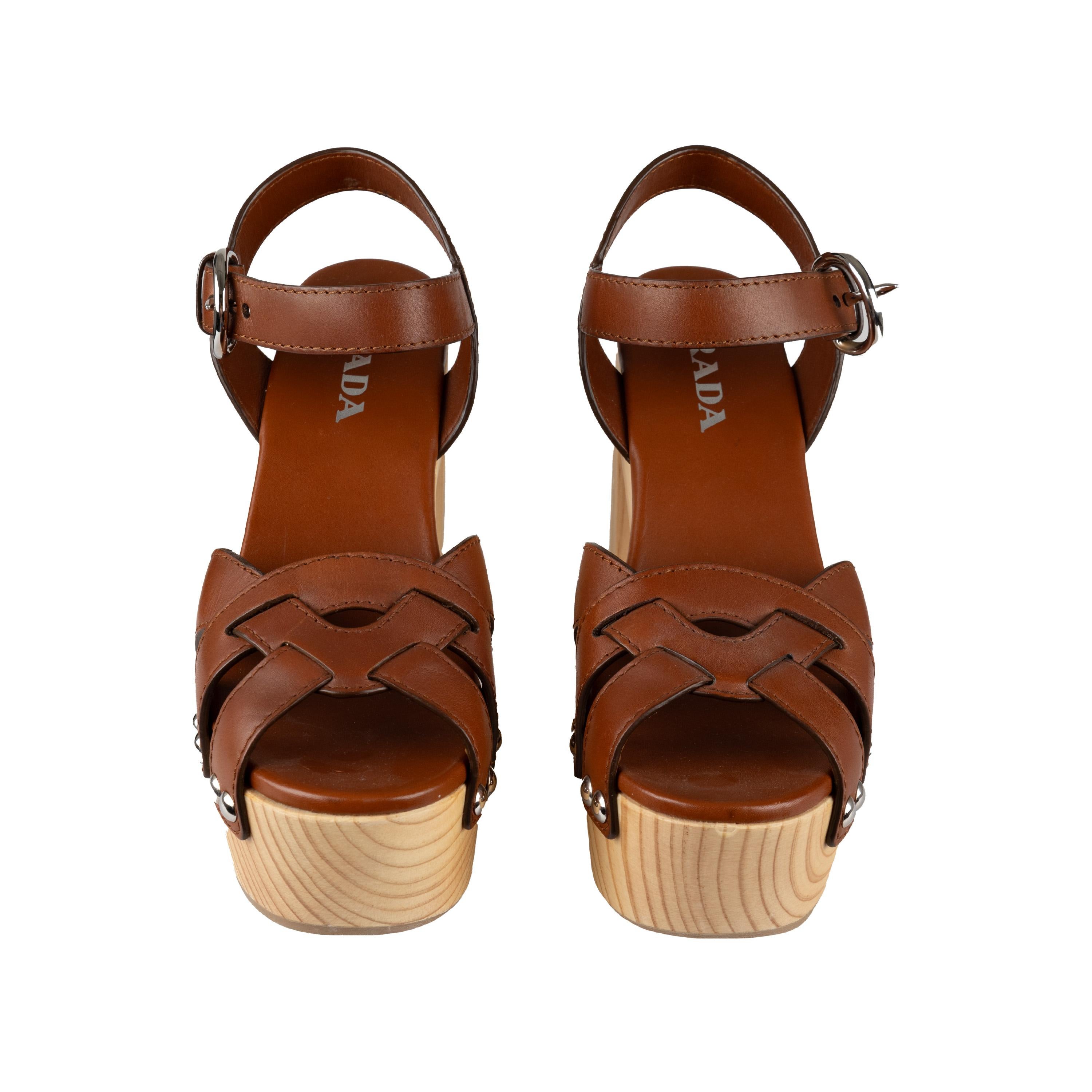 Diese in Italien handgefertigten Prada Plateau-Sandalen sind aus braunem Leder mit geflochtenen Riemen am Vorderblatt gefertigt und mit Nähten und Nieten versehen. Die stabilen Holzplattformen sorgen zusammen mit dem schnallengesicherten,
