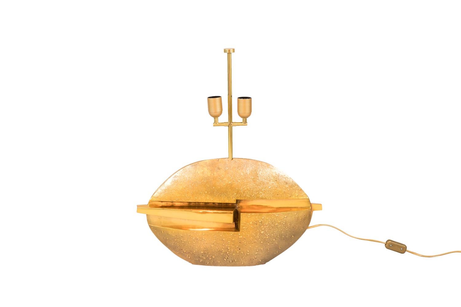 Pragos, signed.
Oval shape lamp in gilt bronze with a granulated surface. Smooth decor of a centered hollow in the whole lamp width and a left projection, on both sides.

Signed Pragos/3.

Work realized in the 1970s.

New and functional