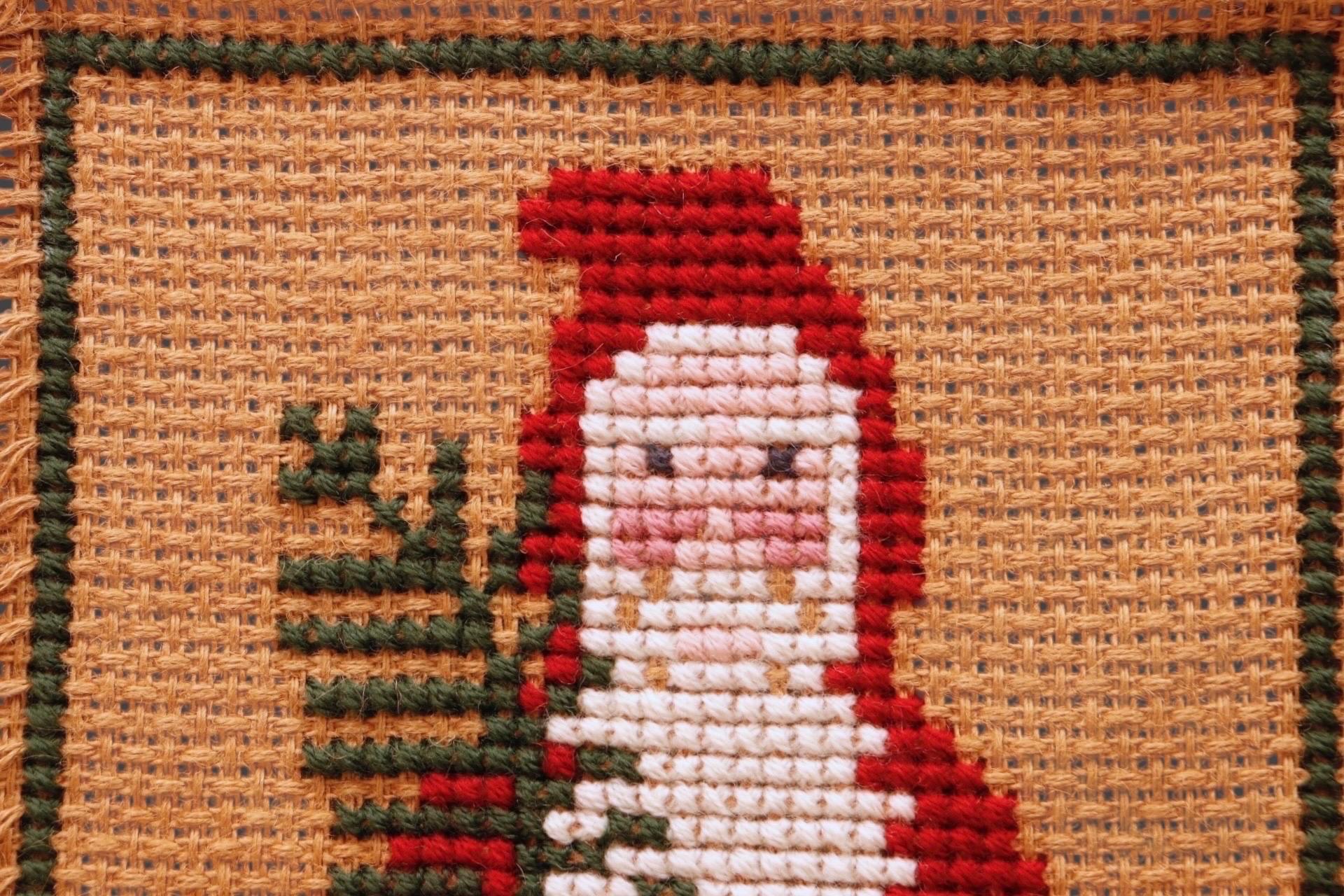 Decoración mural de Papá Noel en punto de cruz sobre arpillera. Un Papá Noel primitivo sostiene una rama, con un peluche en el bolsillo. El diseño, publicado por The Prairie Schooler en 1993, en su 