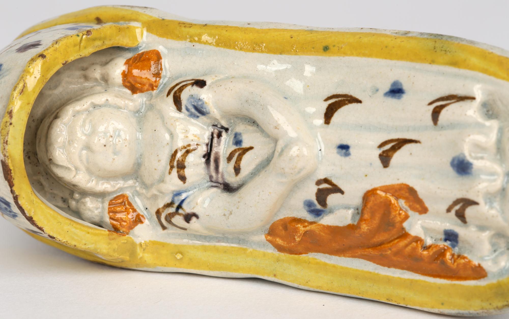 A fine antique Georgian English Prattware pottery cradle containing a sleeping child dating from around 1800. The cradle sits raised on two rockers and modelled as a basket with a hood over one end. The child lies within the cradle asleep. The