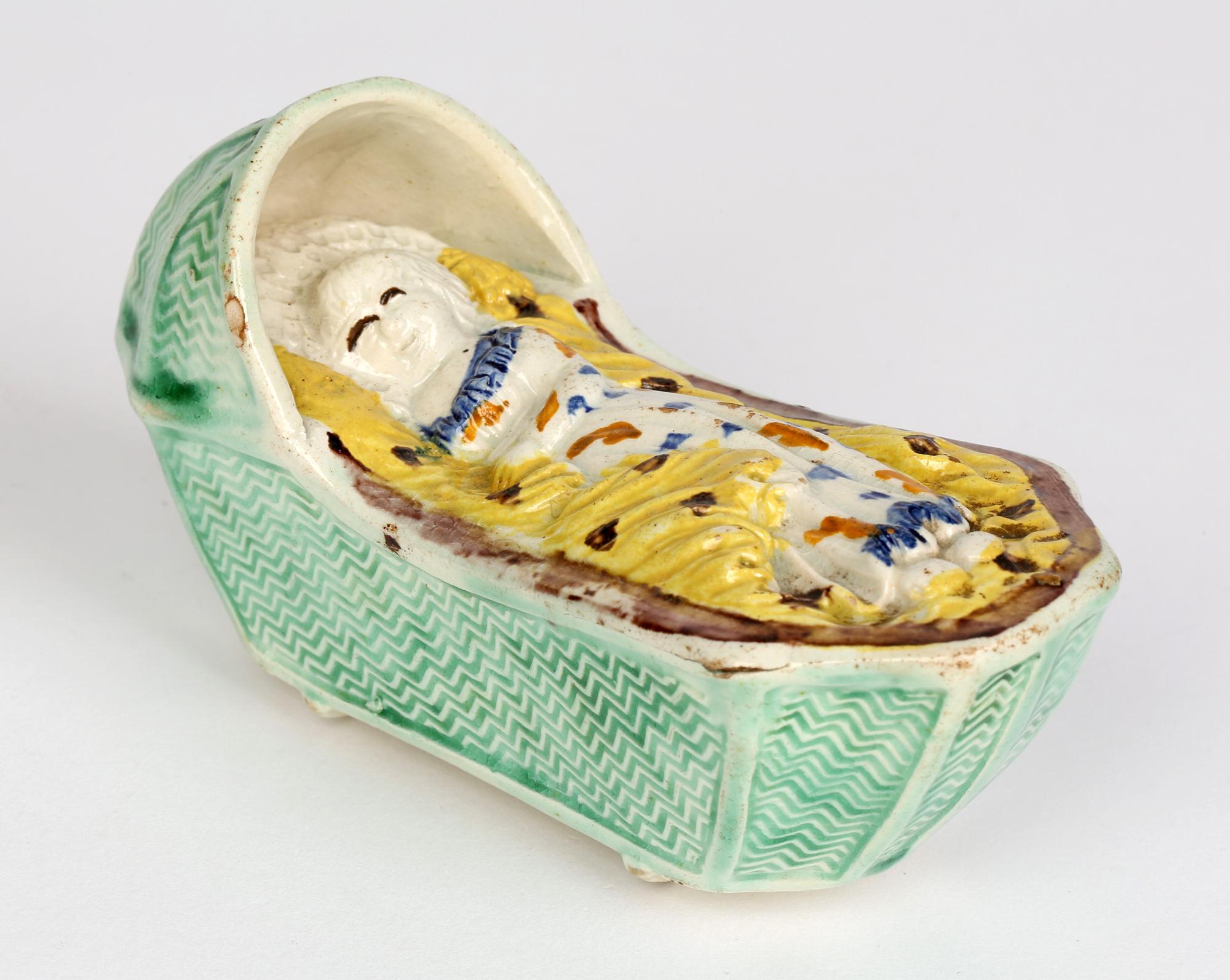 Una bella culla antica in ceramica georgiana inglese Prattware contenente un bambino che dorme, risalente al 1800 circa. La culla è sollevata su due dondoli e modellata come un cesto con un cappuccio su un'estremità. Il bambino giace nella culla