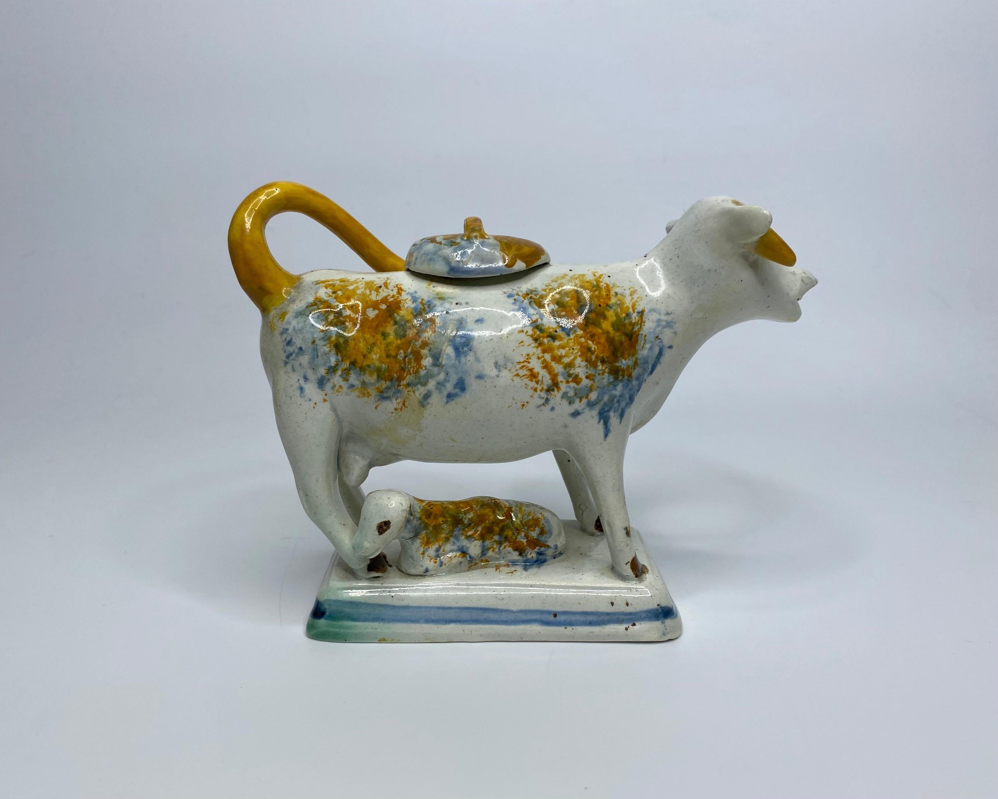English pottery cow creamer, Staffordshire or Yorkshire potteries, c. 1810. The naively potted cow standing upon a rectangular base, with its calf, recumbent at her side.
Painted with Pratt type spots, and colouring to the tail, horns, and cover.