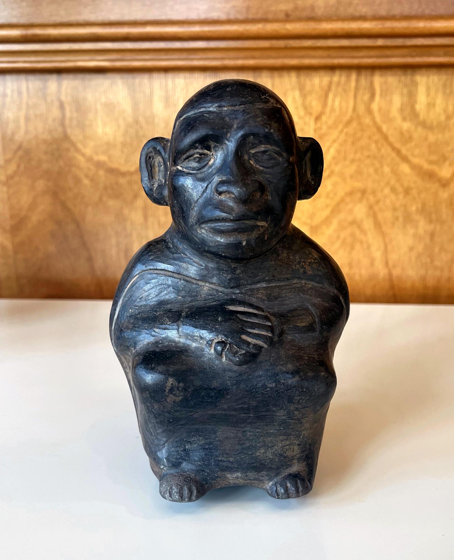 Pequeña vasija estribo figurada de cerámica negra de la cultura Moche circa. 100-500 d.C., situada actualmente en la costa norte de Perú. La olla hueca con asa de estribo se moldeó con sensibilidad y se bruñó la superficie. La figura sentada tiene