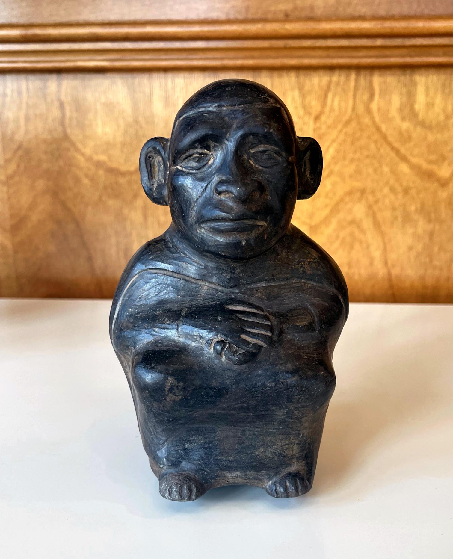 A small black ceramic figural stirrup vessel from Moche culture circa. 100-500 AD, located in nowadays Northern Coast of Peru. The hollow pot with the stirrup handle was sensitively molded and the surface burnished. The seated figure has his arms