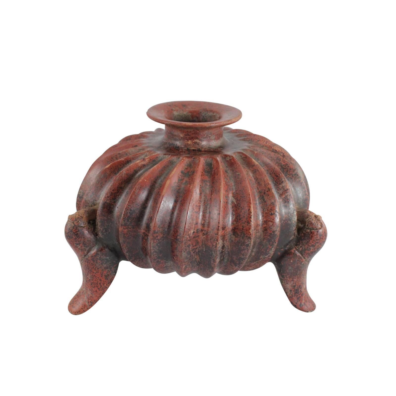 This curious slip-glazed earthenware vessel originated from Pre-Columbian Colima between 200 and 300 BC. The ridging along the sides, called gadrooning, mimics naturalistic forms. Three legs uphold the vessel, which is slipped red and burnished to a