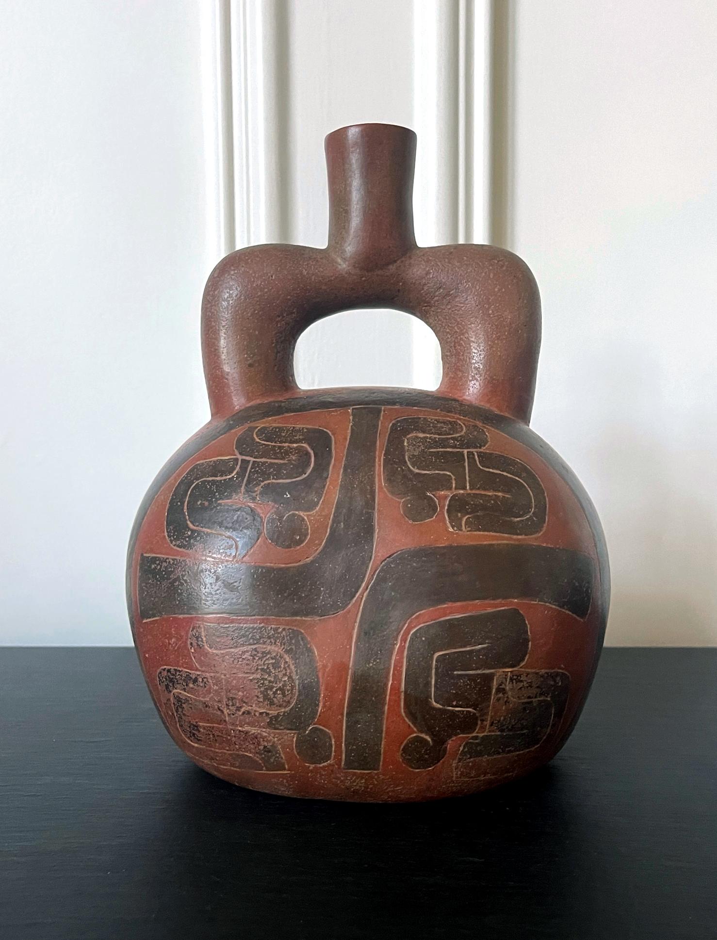 A pre-Columbian ceramic stirrup vessel in Cupisnique style circa 700-500BCE. This ancient vessel features a robust squatted body and a short thick stirrup sprout. Bold geometrical designs in low reliefs colored in black dramatically contrast the red