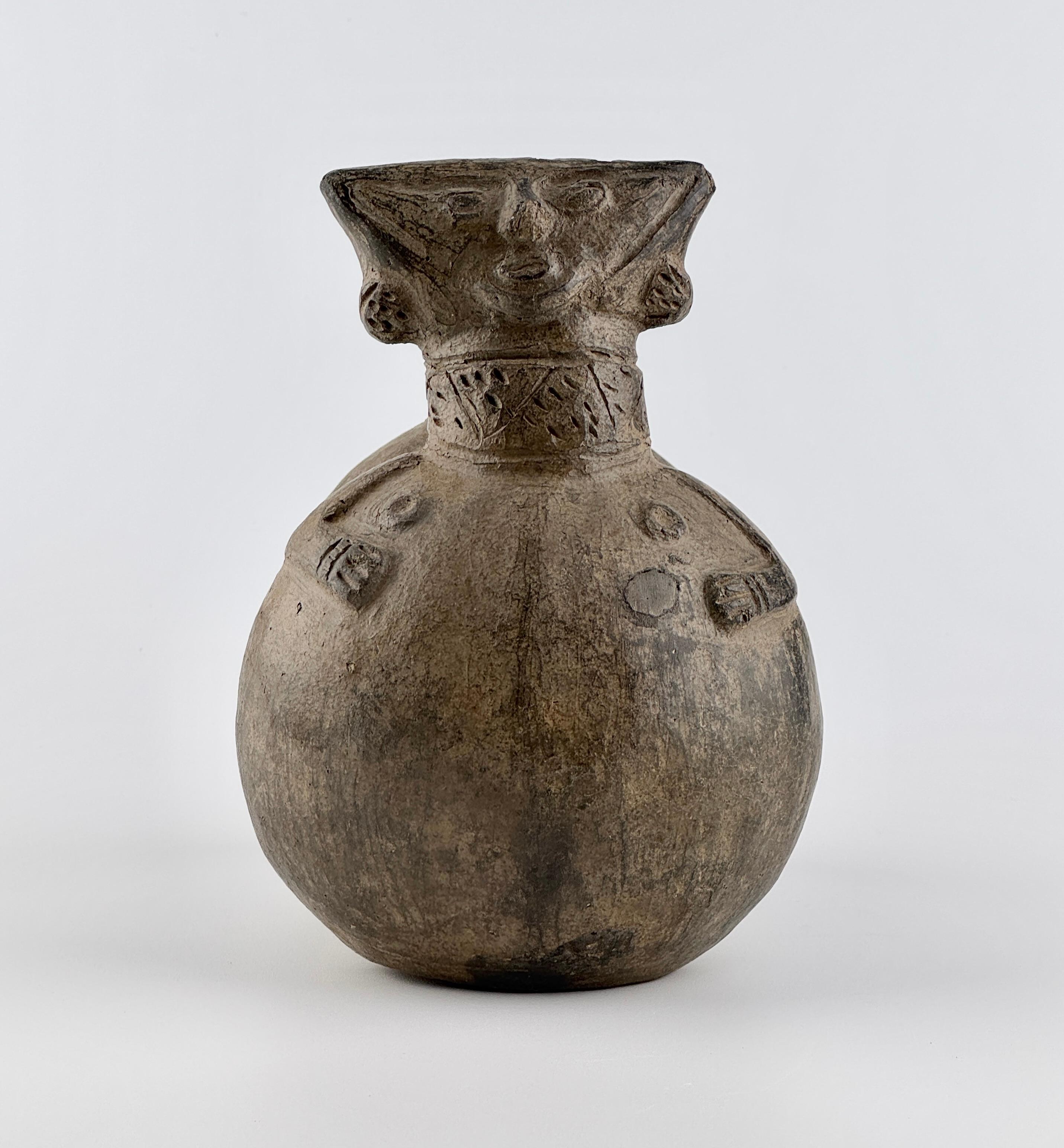 This double-spouted vessel originates from the Peru and is distinguished by its sculptural anthropomorphic design. One of the chambers is shaped as a human figure with a triangular face, deeply incised facial features, and applied ears. The neck is