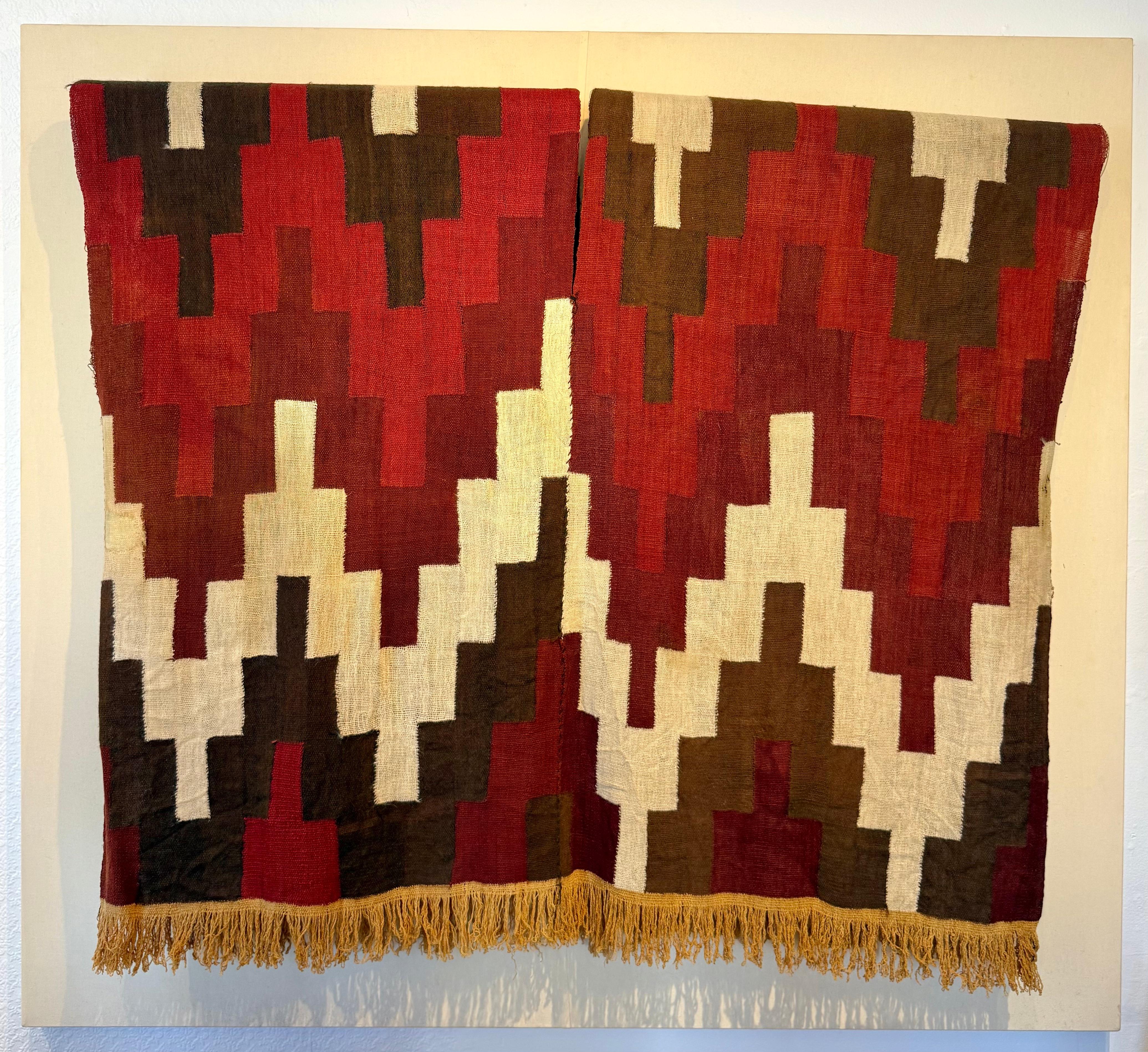 A very rare surviving  Pre-Columbian Nazca Stepped Textile Tunic, Nazca Peru, 200-600 AD.  Museum mounted on a linen covered panel. Impressive piece overall.

Stepped design and fringe in yellow ocher, burgundy and brown.  Excellent conditoin for