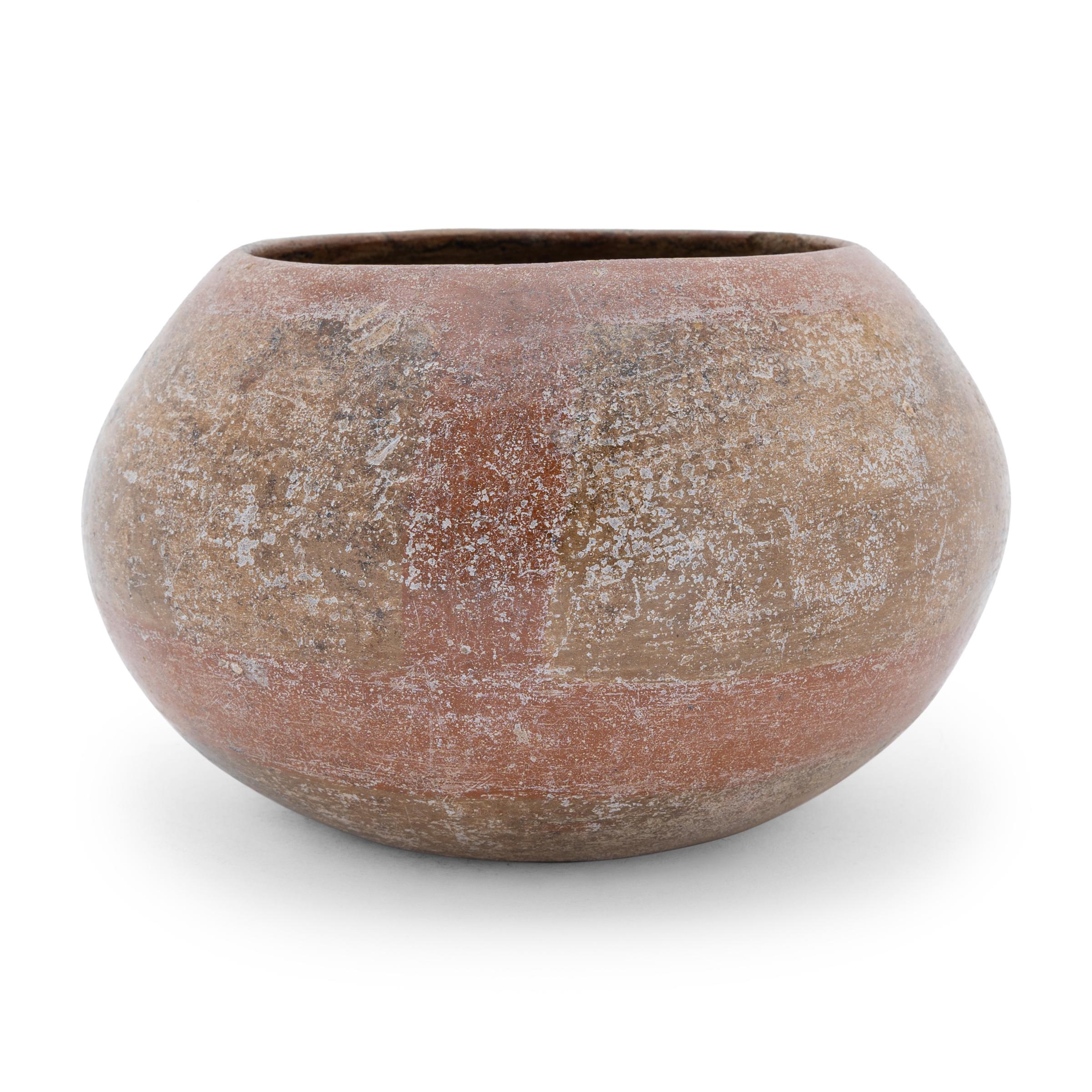 With a worn finish, this round orangeware bowl is a wonderful example of Pre-Columbian pottery. The delicate bowl has a squat form and tapered lip. Painted with stokes of red slip and speckled with white etching, this jar has an aged beauty. The