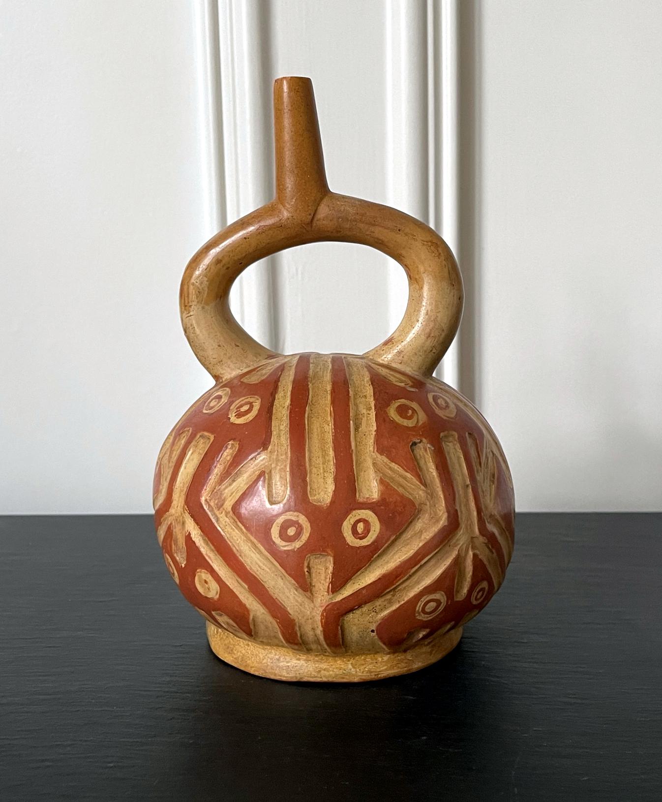 A ceramic stirrup vessel pot from late Moche culture circa 500-700AD in nowadays Northern Coast of Peru. 
The hollow vessel features a rounded body and a stirrup handle and sprout, supported by a short foot ring. The highly stylized surface is