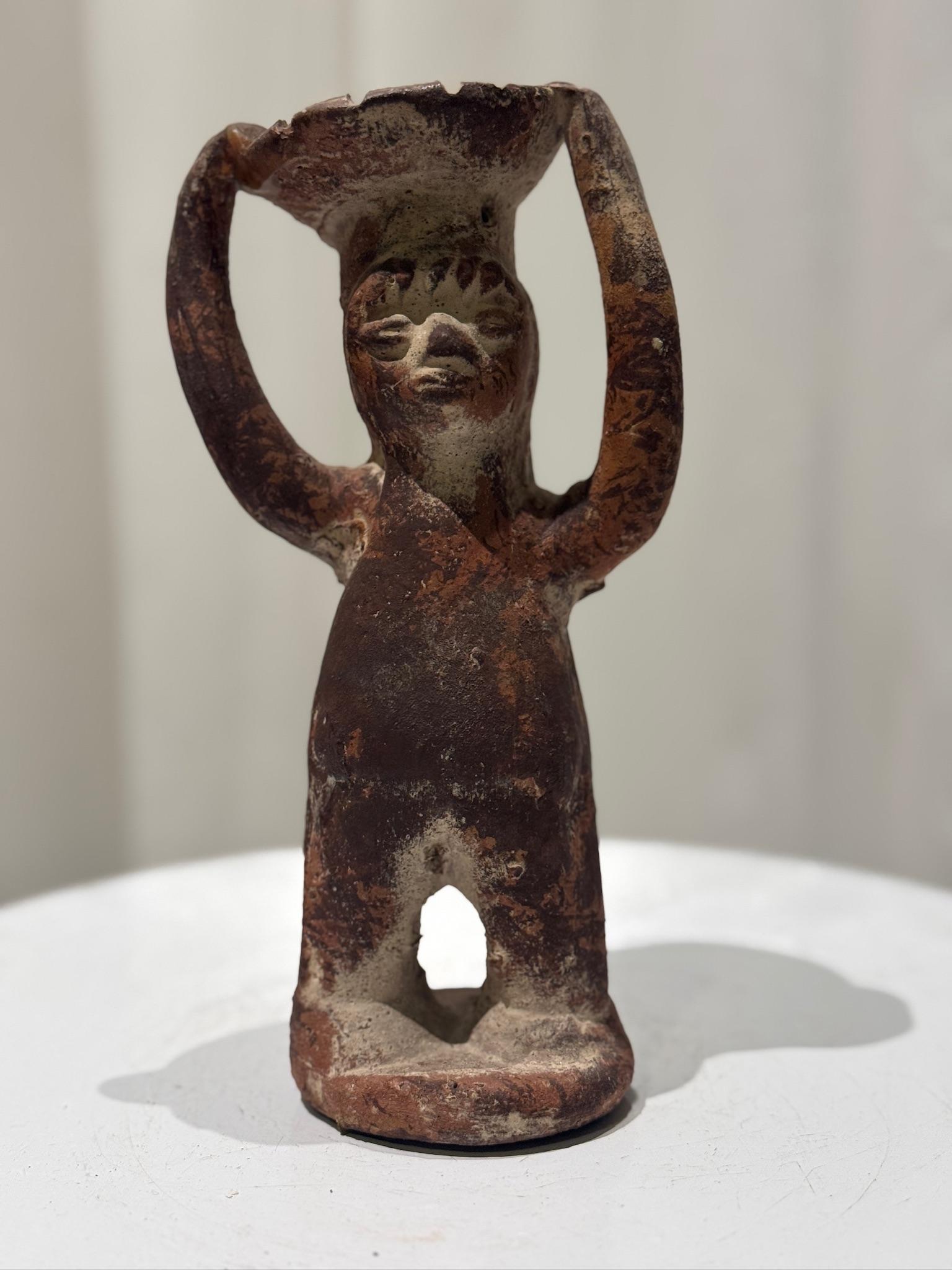 This Pre-Columbian style terracotta sculpture is a remarkable representation of early Mesoamerican artistic traditions, handcrafted in a primitive yet evocative manner. The figure, depicted with both arms raised, appears to be supporting a dish or