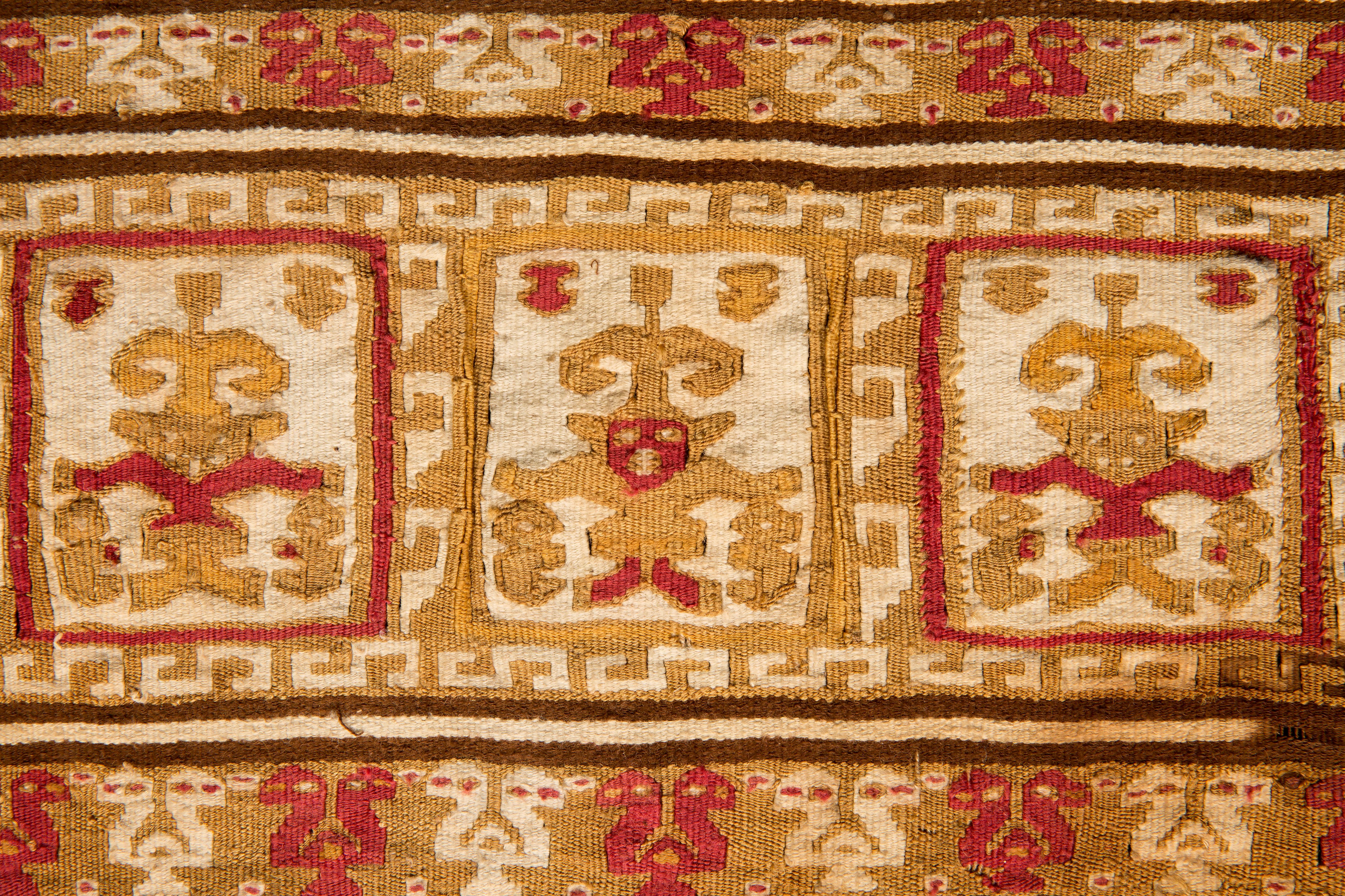 Pre-Columbian ornamental banner with fringes composed of warriors/divinities holding figures in their hands, surrounded by geometric frets. Two-headed birds in alternating colors decorate the top and bottom of the banner. This one-of-a-kind tapestry