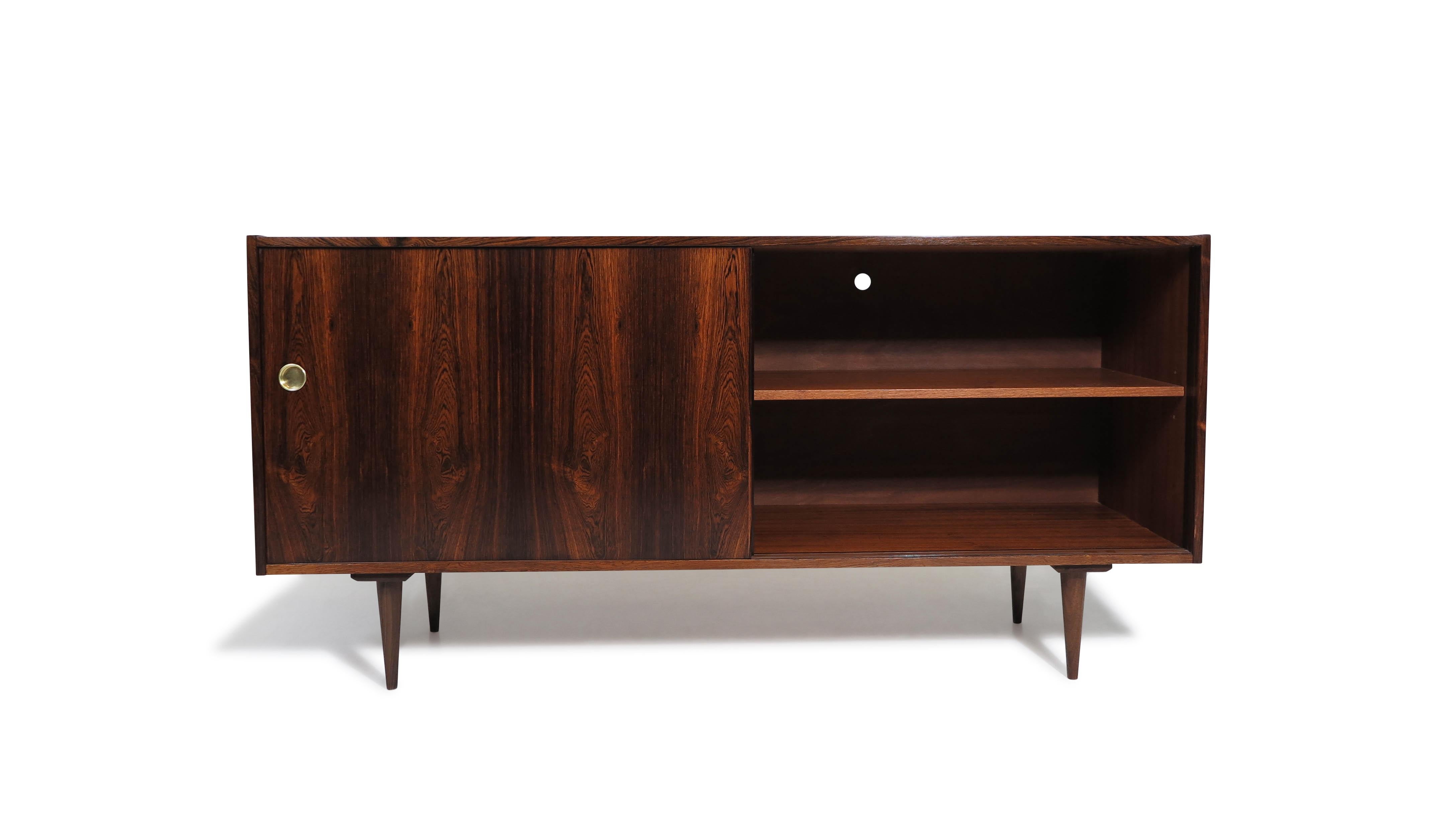 Mid-century Scandinavian sideboard crafted from Brazilian rosewood, featuring book-matched grain across a pair of sliding doors with recessed brass pulls. Raised on round tapered legs, the cabinet interior is made of Cuban mahogany and includes