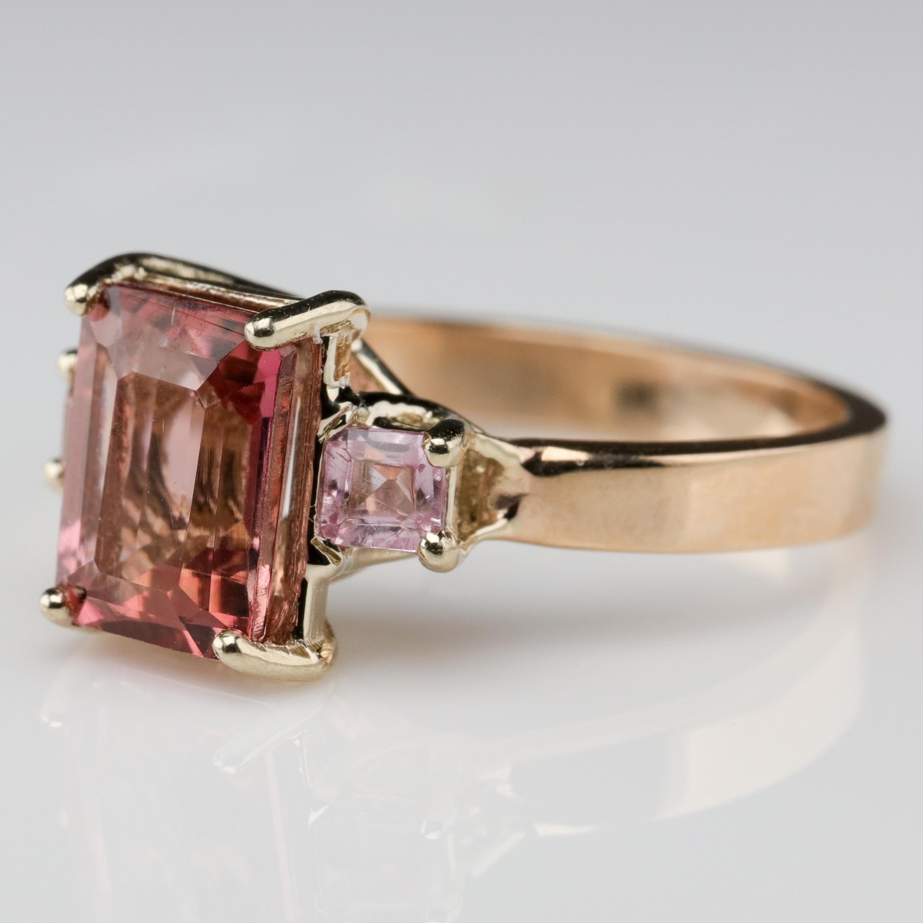 This ring was hand-made by an old-school jeweler in Pennsylvania and features a peachy-purplish-pink emerald-cut precious topaz stone from Brazil that measures approximately 8.14 mm x 6.06 and weighs approximately 1.85 carats. On either side are two
