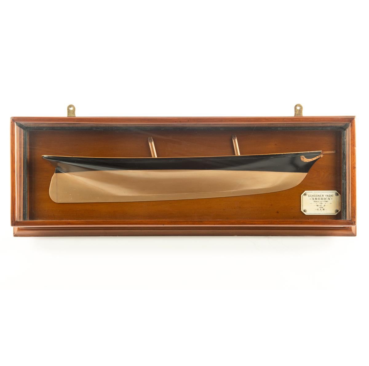 This cased presentation half hull model is of the first winner of the America’s Cup, the schooner America. The copper coloured bottom is painted with black topsides and has a finely carved garland around the bow. There are two stump masts, a caprail