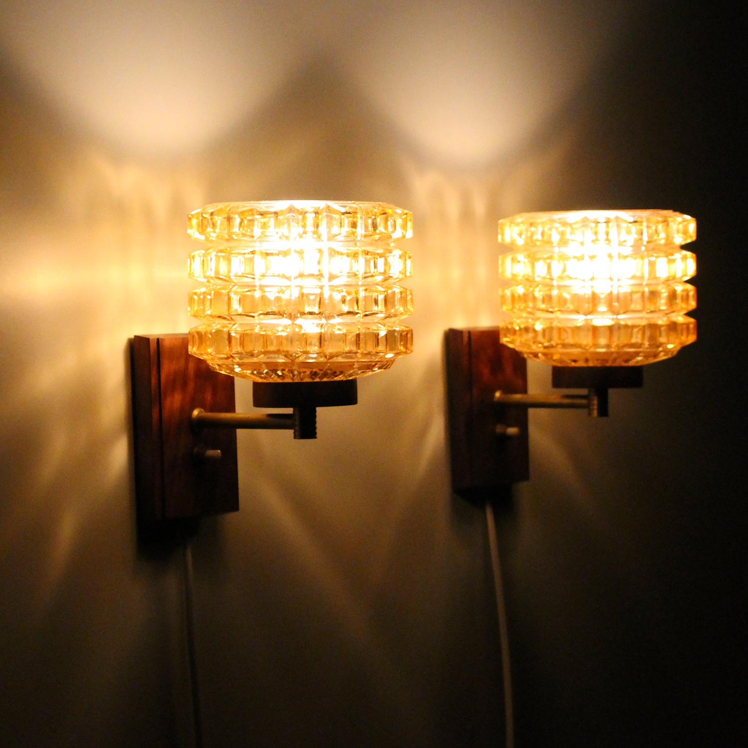 Pressed glass and rosewood wall lamps, 1950s Scandinavian midcentury wall lights with amber glass, brass and rosewood in very good vintage condition.

This beautiful pair of wall lights, each made up of a thick pressed glass shade, a brass arm and a