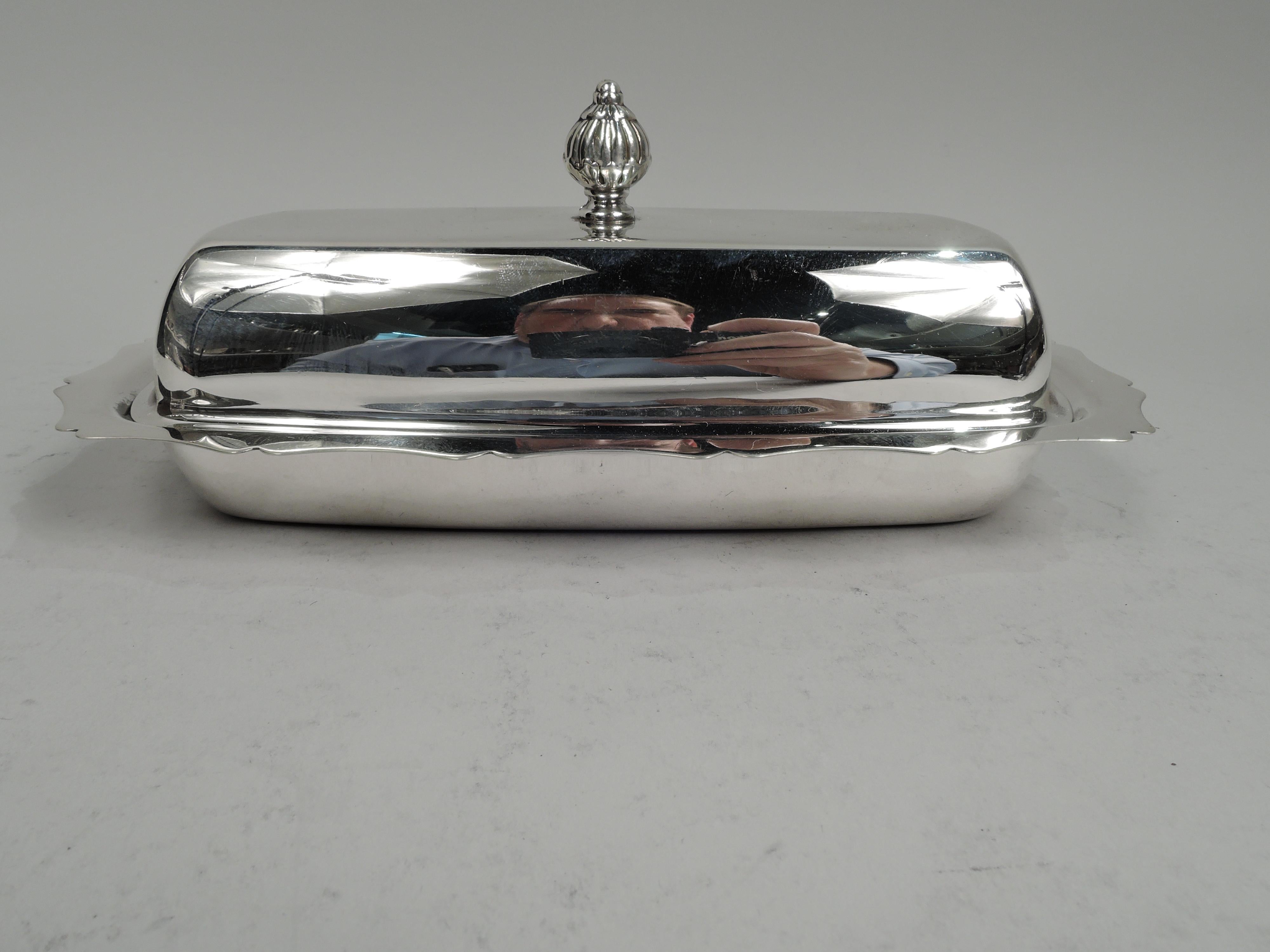 Pretty sterling silver butter dish. Made by Fisher Silversmiths, Inc. in Jersey City, ca 1950. Long and rectilinear well with curved ends and flat scrolled rim. Cover raised with lobed finial. With clear glass liner with radiating flutes. Fully