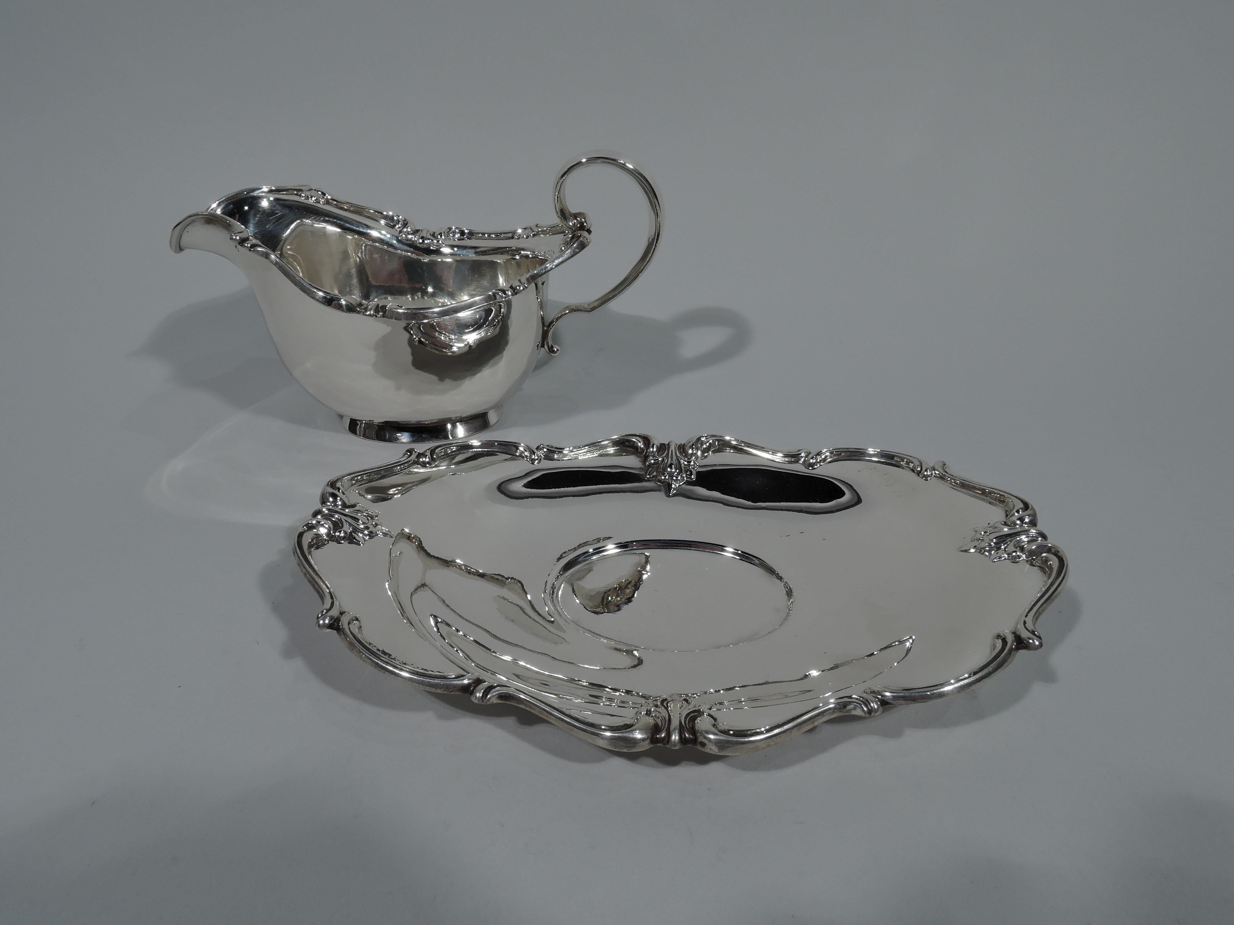 Pretty sterling silver gravy boat. Made by Frank M. Whiting in North Attleboro, Mass., circa 1940. Boat: Curved sides, high looping scroll handle, and short splayed foot. Ruffly asymmetrical rim with scrolls and leaves. Stand: Curved with foot ring.