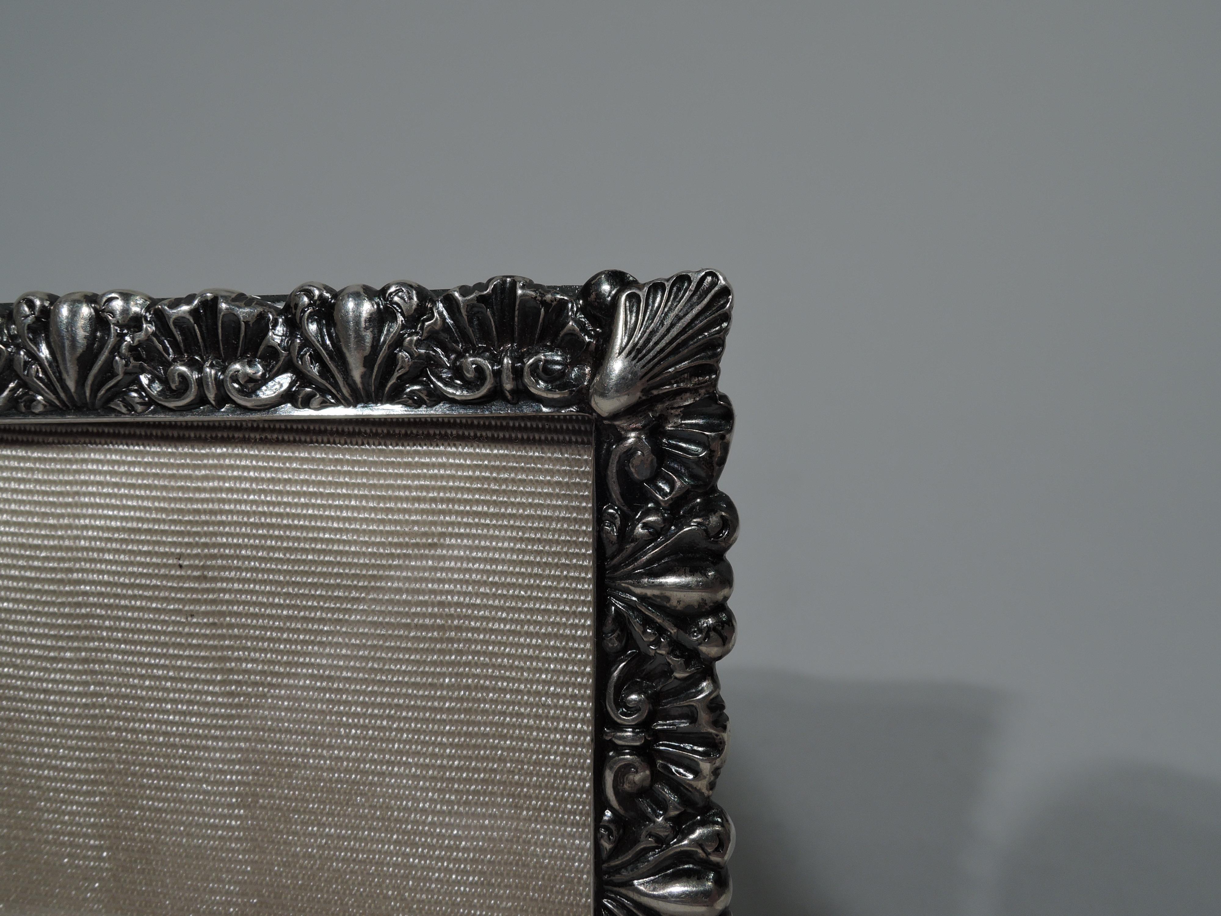 Pretty sterling silver picture frame. Made by Bigelow, Kennard & Co. in Boston, ca 1900. Rectangular window surrounded by alternating tooled shells and leaves. With glass, silk lining, and hinged silver wire support. Fully marked. 

Dimensions: