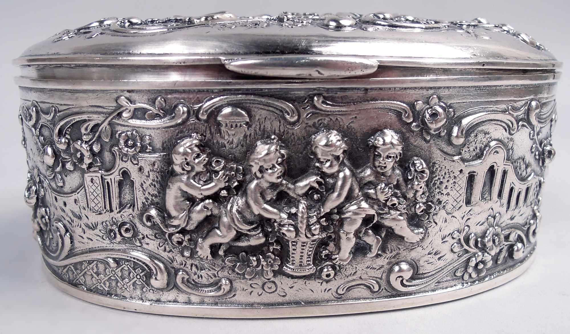 Hübsche deutsche Rokoko-Silberdose, um 1910. Oval mit aufklappbarem Deckel. Ziselierte und gravierte verschnörkelte Rahmen mit Gruppen von Putten, die sich um einen Korb scharen; im Hintergrund klassische Architektur. Innen vergoldet. Stempel: