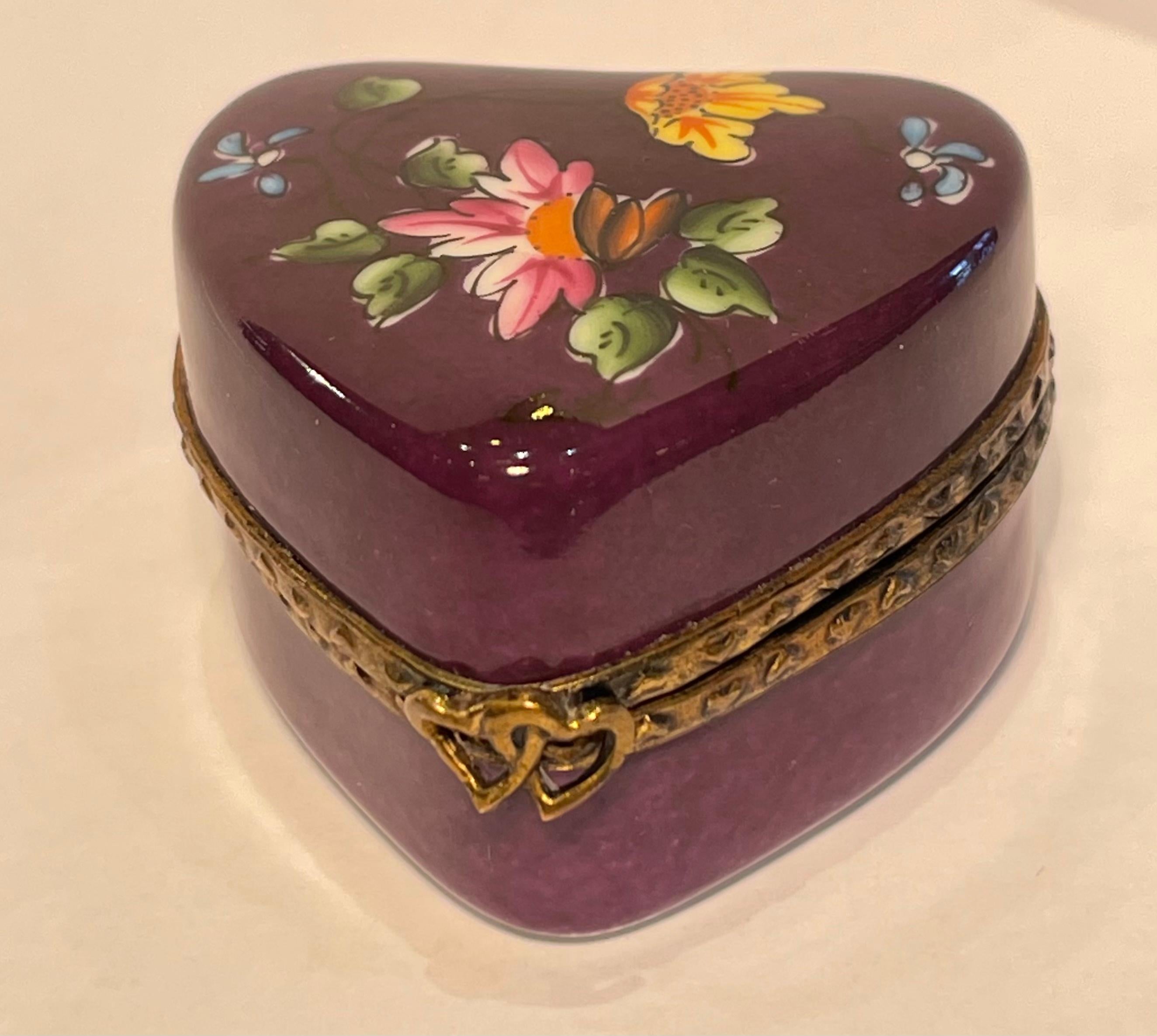 Collectible handmade and hand painted Limoges porcelain miniature heart shaped trinket box features a rich deep purple background with a beautiful hand painted polychrome floral motif on the top. The fittings are a richly patterned antique gold gilt