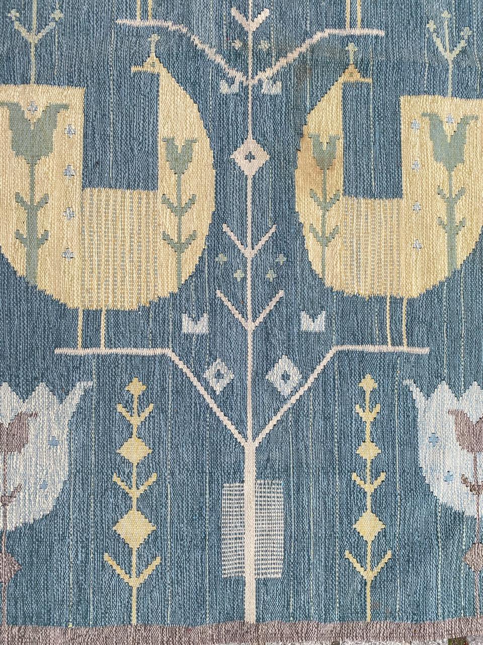 This woven woollen tapestry features a design of stylised birds and flowers. It is woven in a heavyweight wool using mainly nature-inspired shades of sky blue, yellow and ecru highlighting the flowers and birds. It has the designer's initials NE in