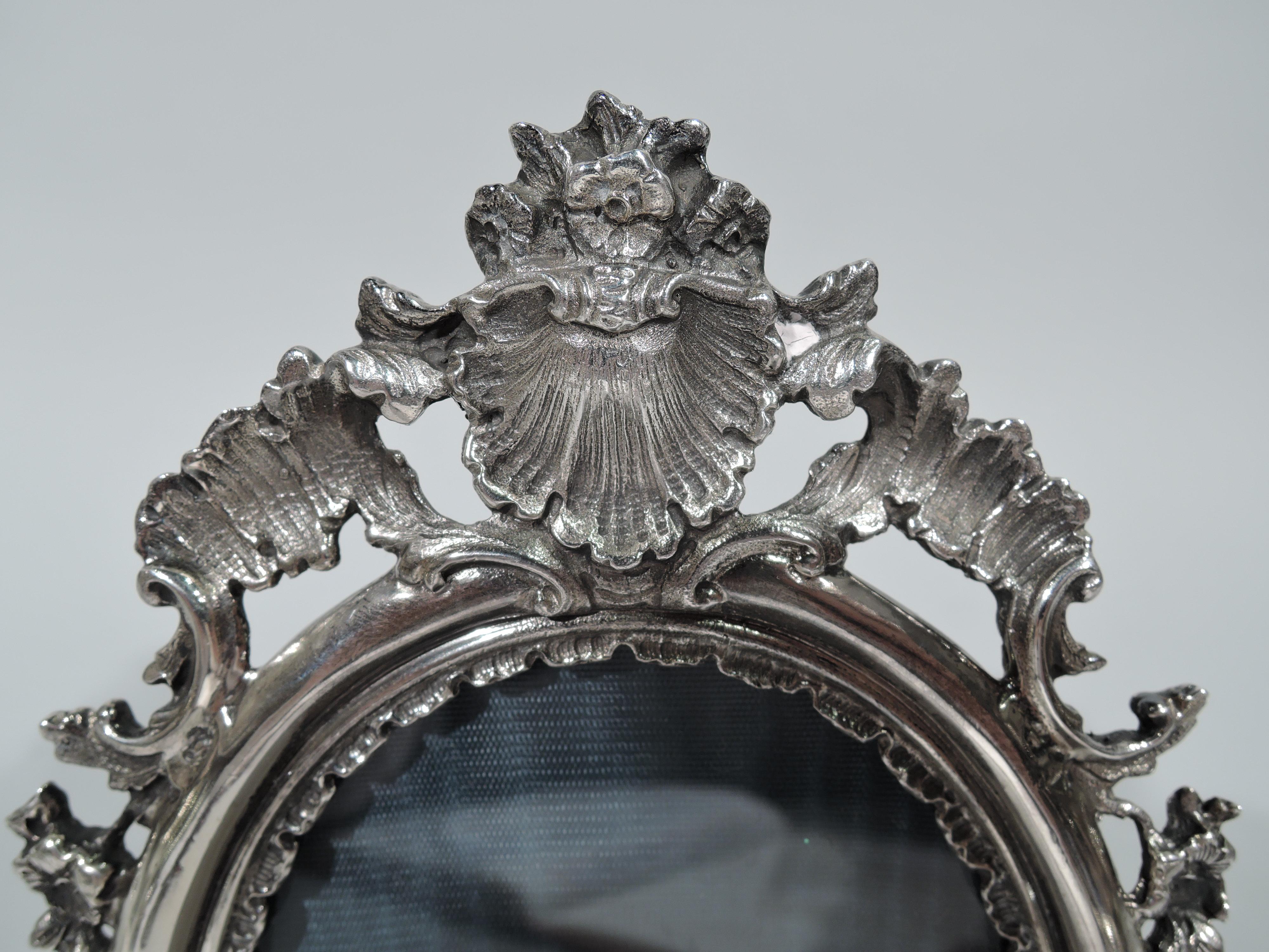 Pretty Rococo sterling silver picture frame. Oval window with molded surround and interior leaf border. Open shells, leaves, and flowers applied to top and bottom. With glass, silk lining, and leather back and hinged support. Post-1967 Italian