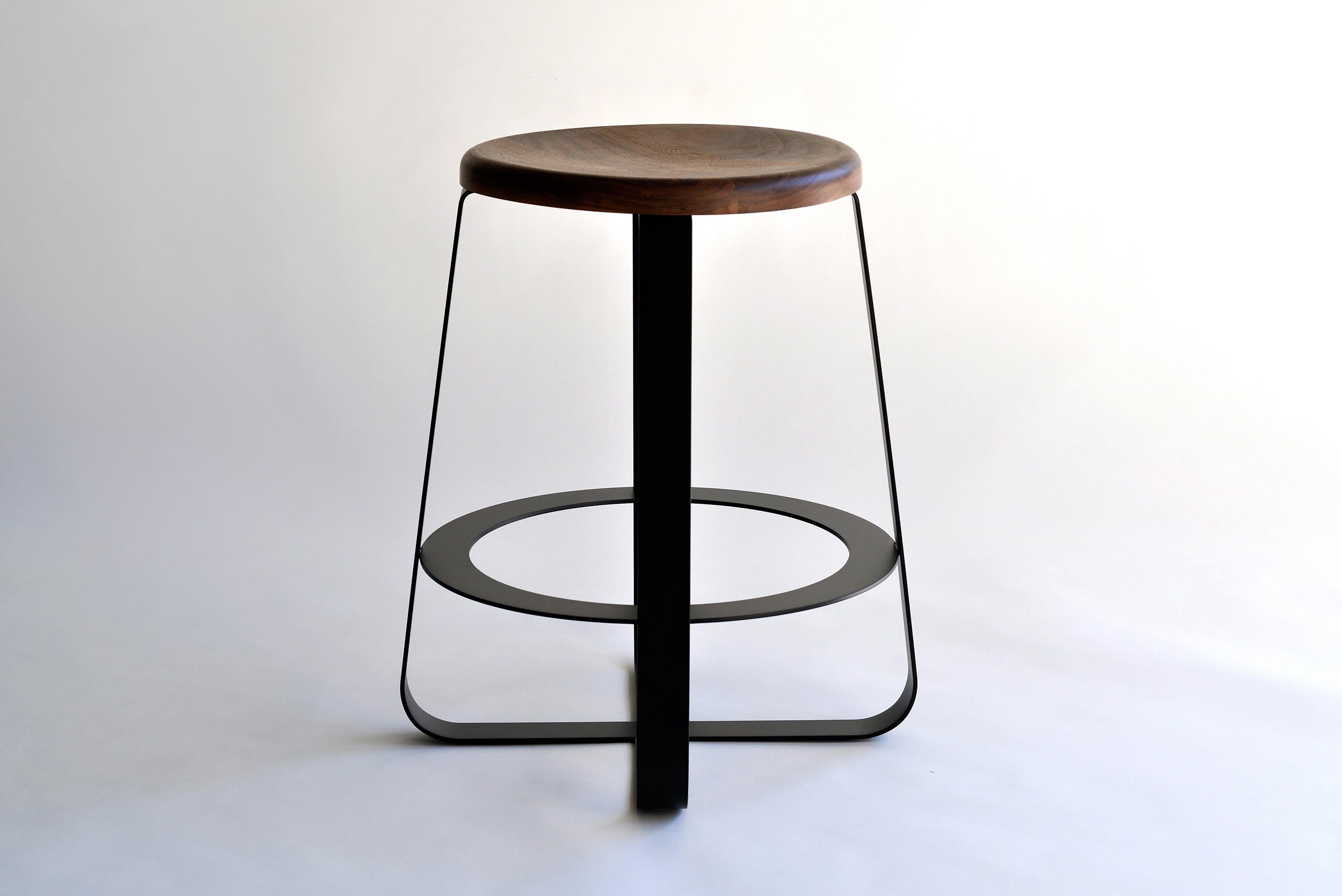 Primi Counter Stool by Phase Design
Dimensions: Ø 54.6 x H 62.2 cm. 
Materials: Powder-coated steel and walnut.

Solid steel flat bar available in a flat black or white powder coat finish with carved walnut, white ash, or upholstered top. Powder