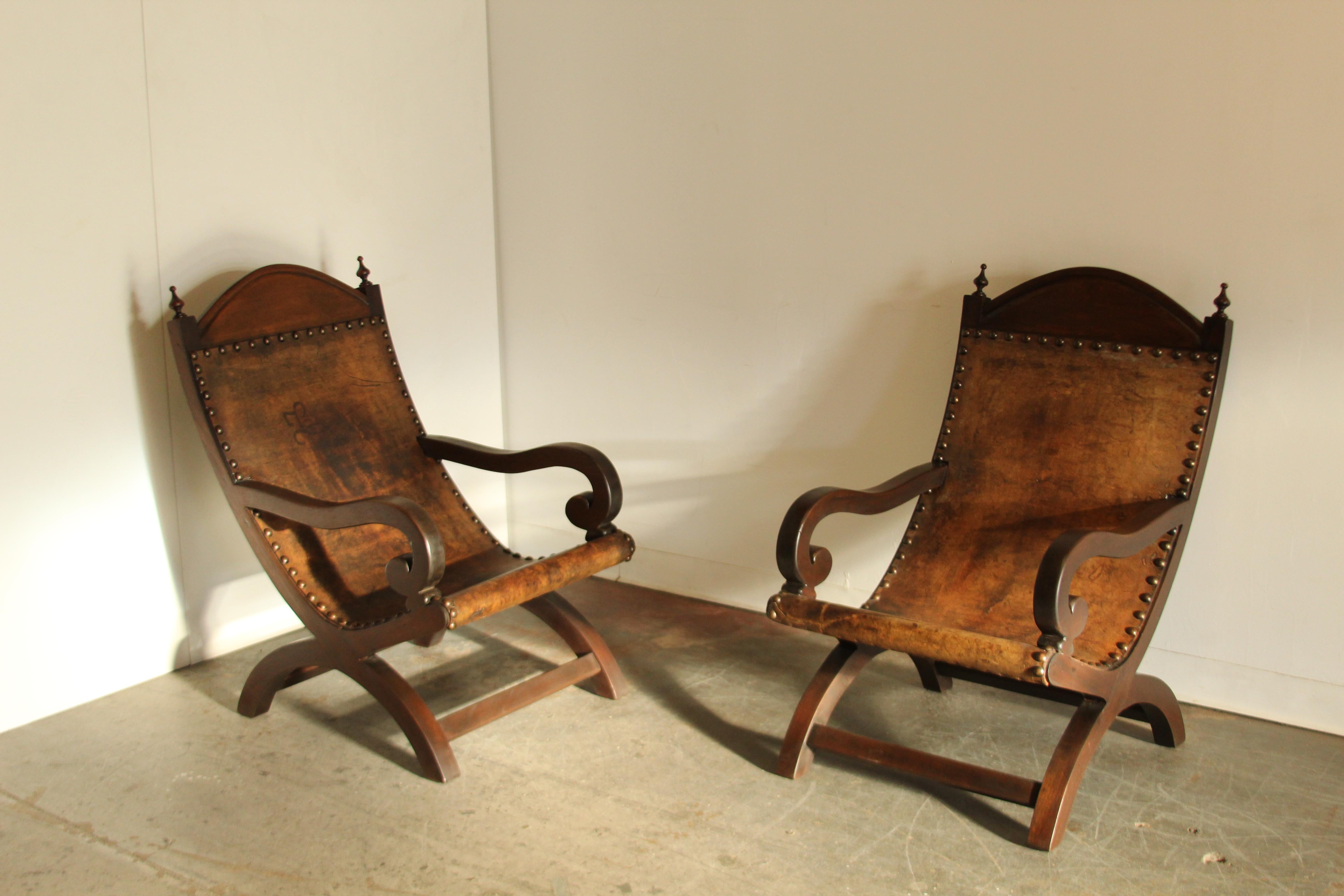 A wonderful pair of leather butaque chairs made in Mexico in the 1970s. Delightful rustic, primitive aesthetic. Solid wood frames with classically curled arms, scoop form, and the tops adorned with ornate finials. Thick, distressed leather cowhide