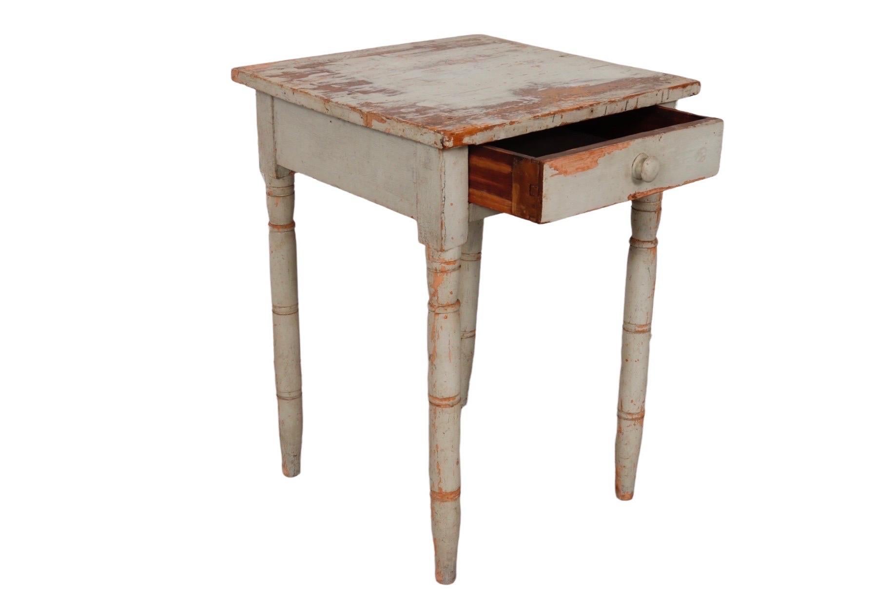 A primitive painted side table. A plank wood top and lightly turned legs house a single hand dovetailed drawer that opens with a knob handle. The light sage colored paint finish is highly distressed with craquelure throughout and some loss of paint.