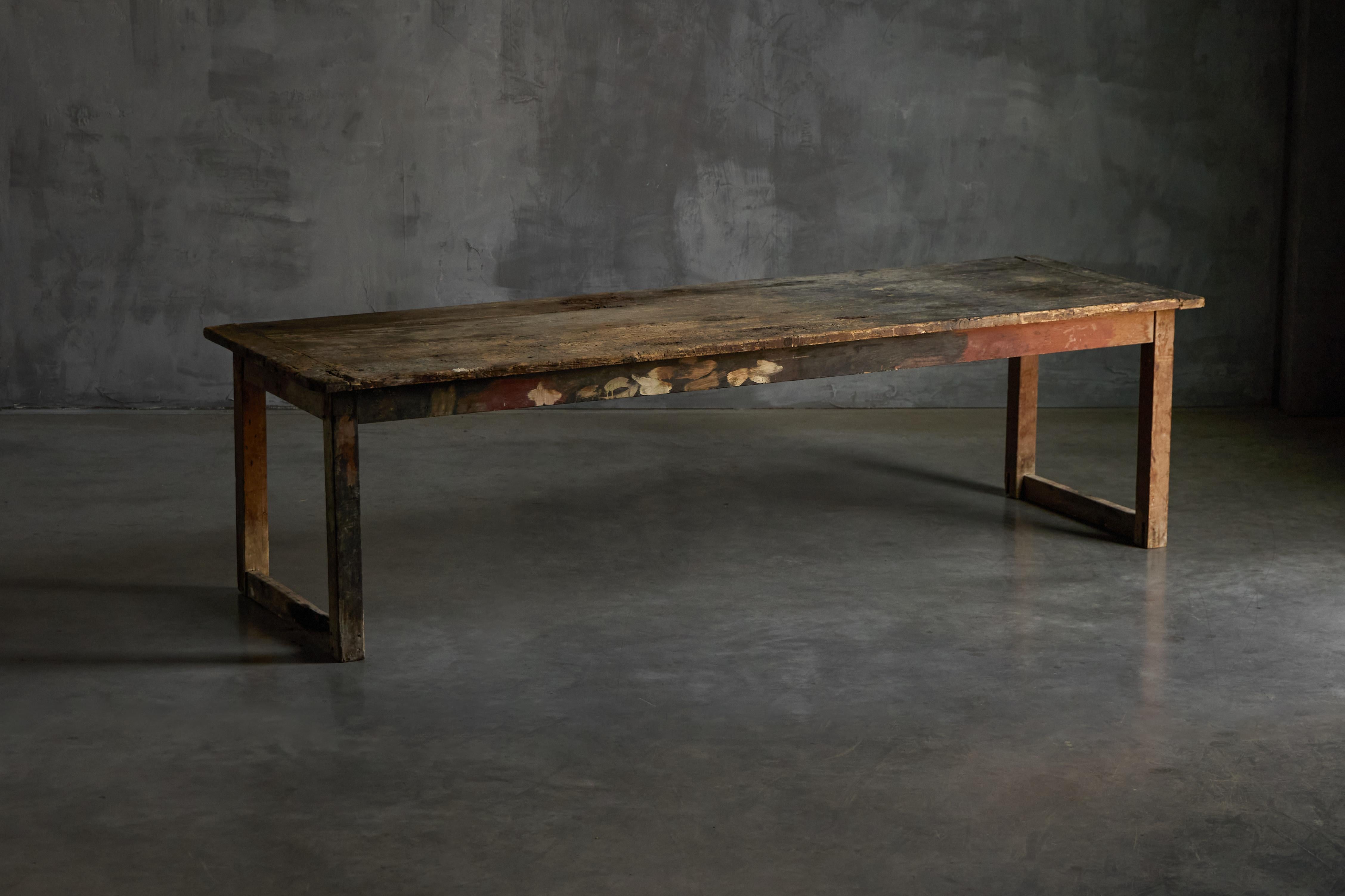 This rustic painter's table dates back to the early 1900s and bears the raw, expressive patina of decades of creative use. Crafted in solid wood, the surface is worn and richly textured with traces of pigments, stains, and brushstrokes that speak to
