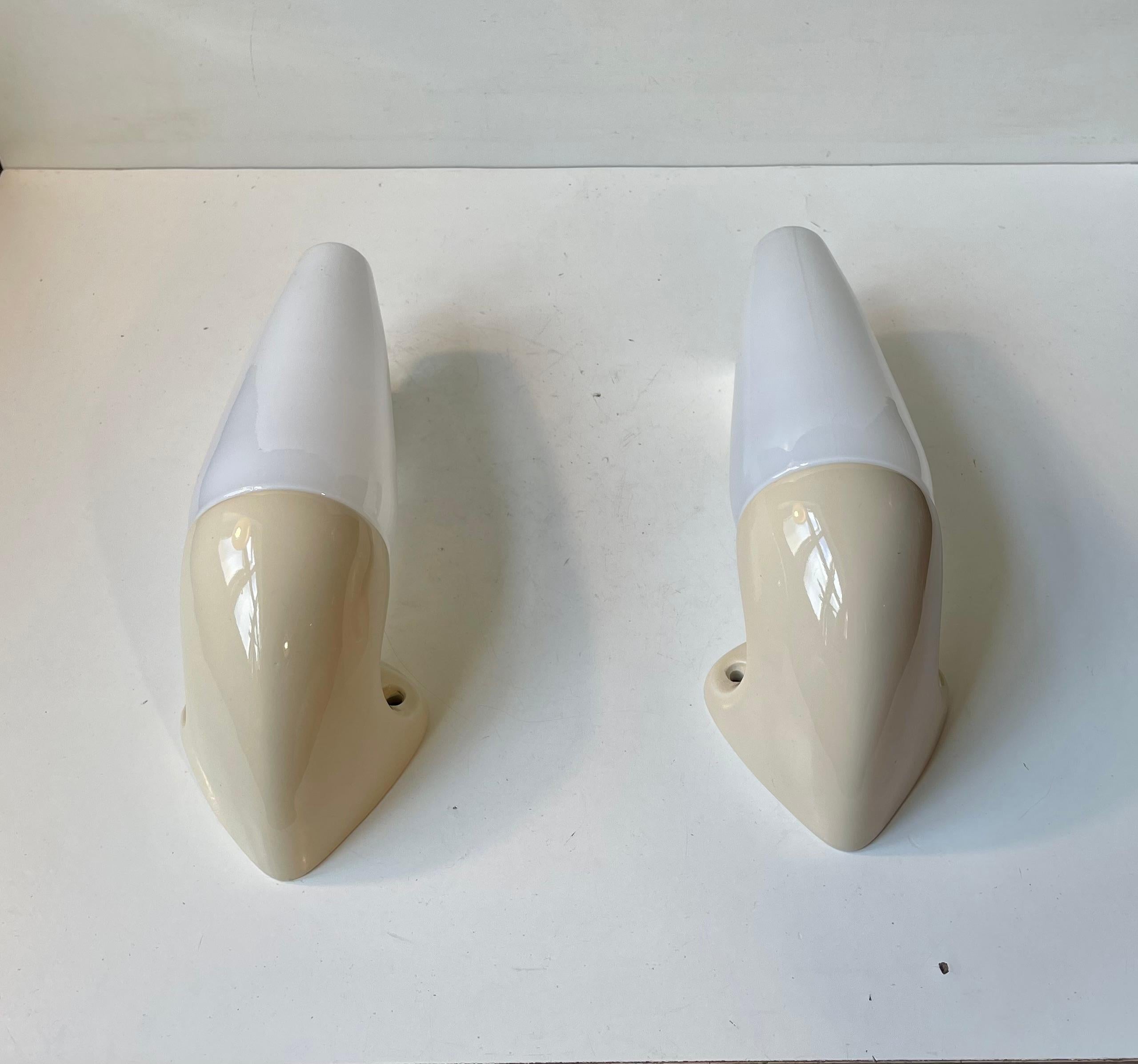 A pair of sconces suitable as bathroom or outdoor lighting. They were designed by the Swedish Prince Sigvard Bernadotte for the swedish company Ifö during the 1960s. Pastel cream glazed porcelain mounts with white opaline glass shading. 2 way mount
