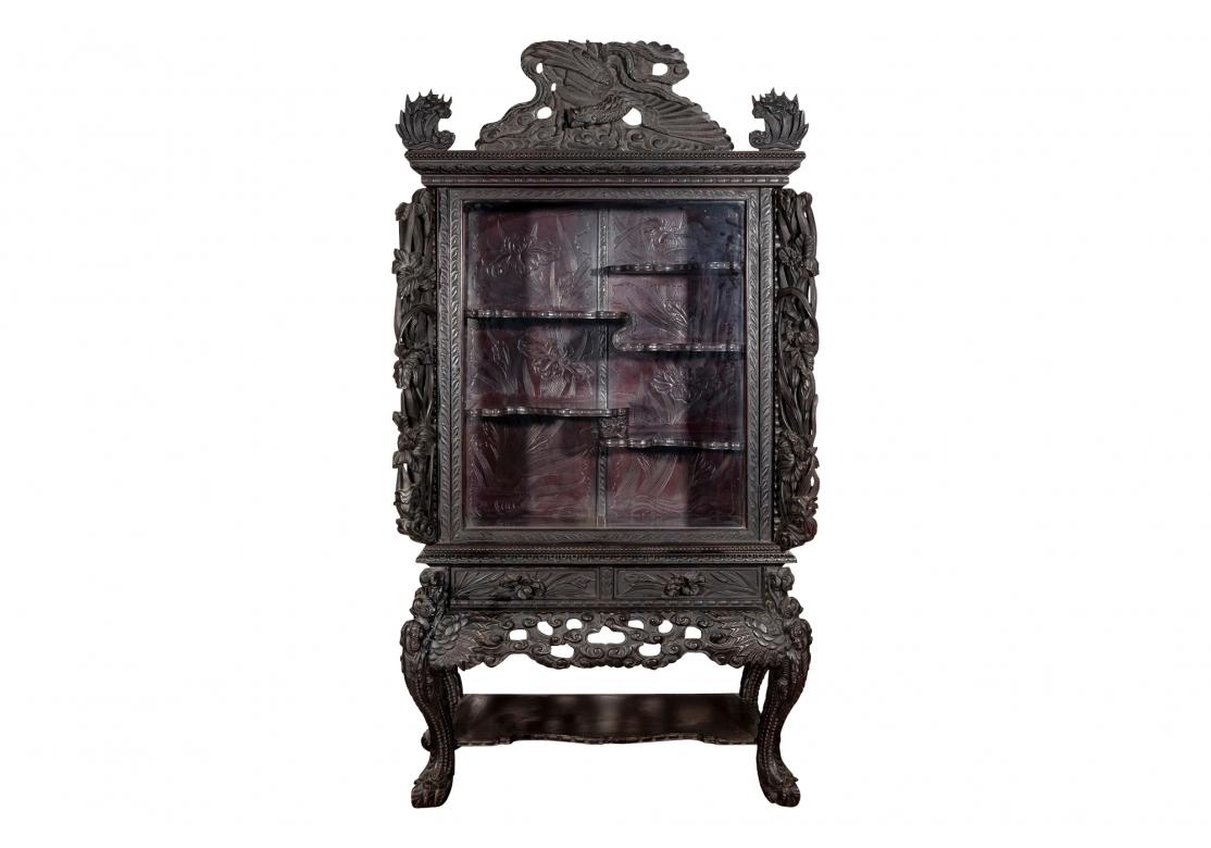 An elegant Asian display cabinet, profusely carved throughout with glass door with carved trim and surround that reveals two long and one short shaped interior shelves. The interior back of the cabinet carved throughout with floral and foliate
