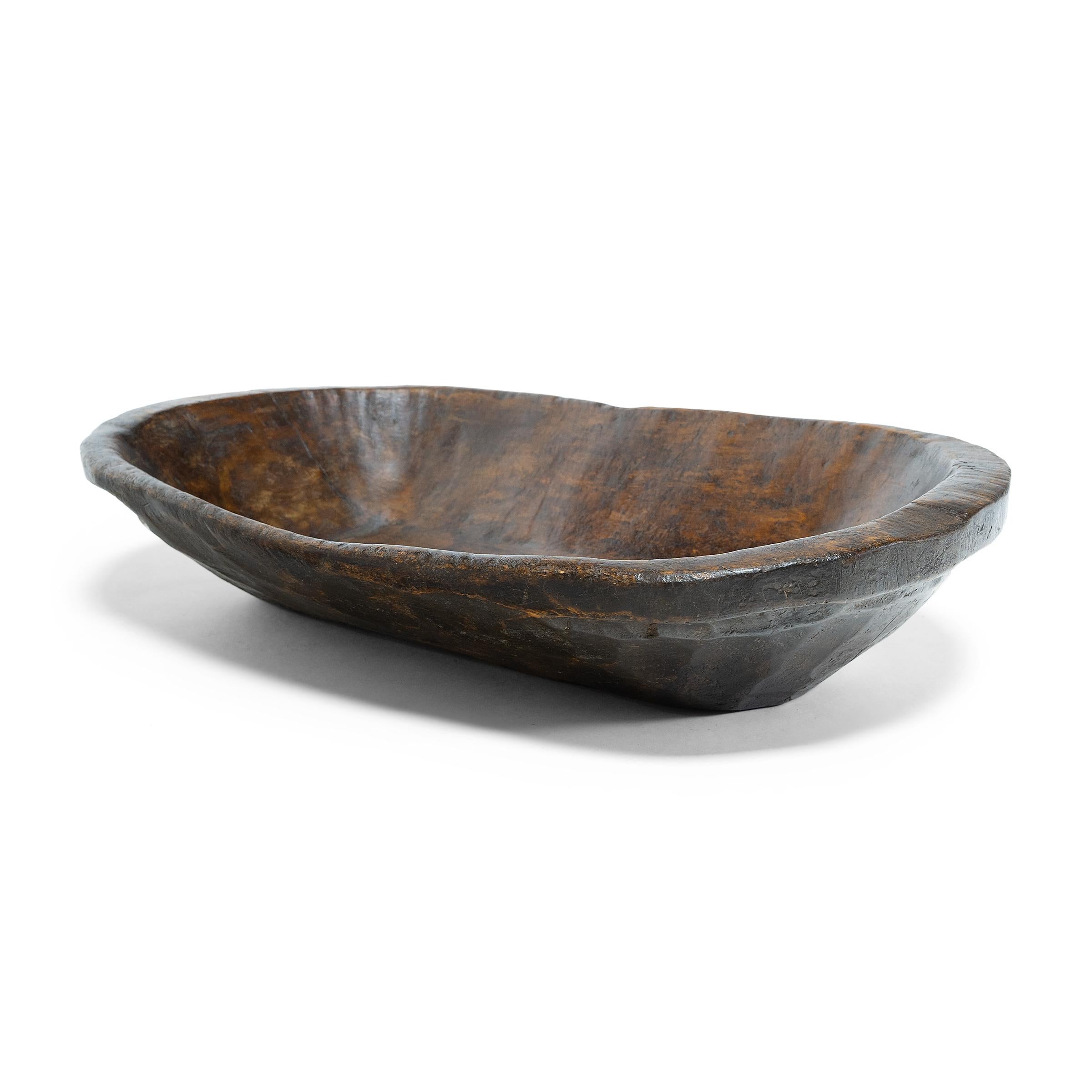 This wooden tray from northern China charms with rustic texture and asymmetric form. Hand-carved from a single block of wood, the tray has an oval shape with rounded corners and tapered sides that flatten at the rim. Years of use have imparted a