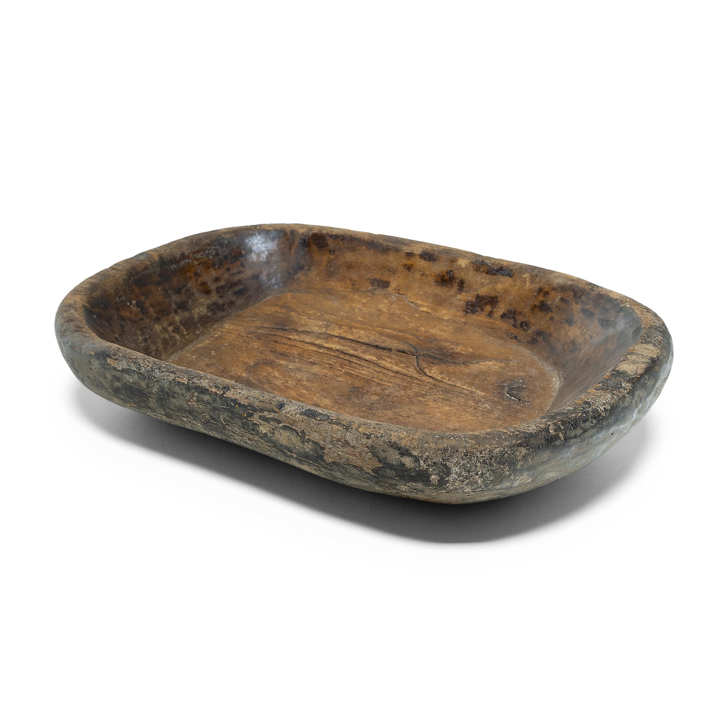 This wooden tray from northern China charms with rustic texture and asymmetric form. Hand-carved from a single block of wood, the tray has an oval shape with rounded corners and short, tapered sides. Years of use have weathered the original lacquer